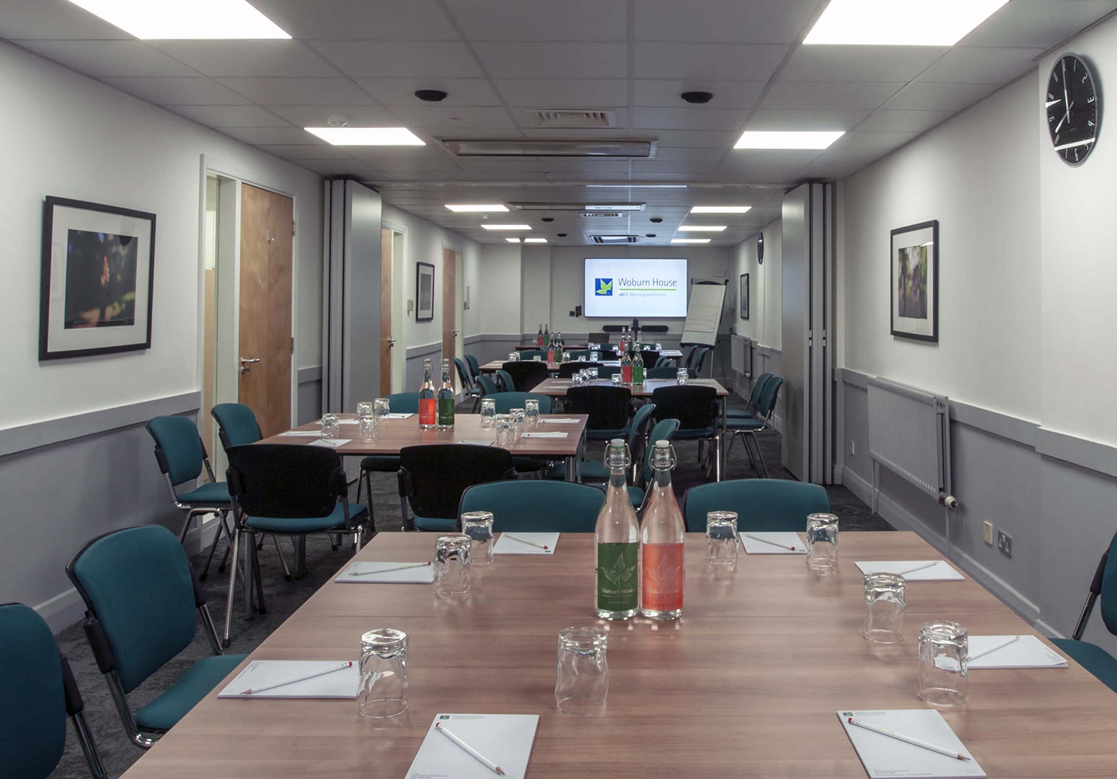 A conference room is set up with multiple tables arranged in a U-shape, equipped with chairs, notebooks, and water bottles, and features a screen displaying the "Walton House" logo.