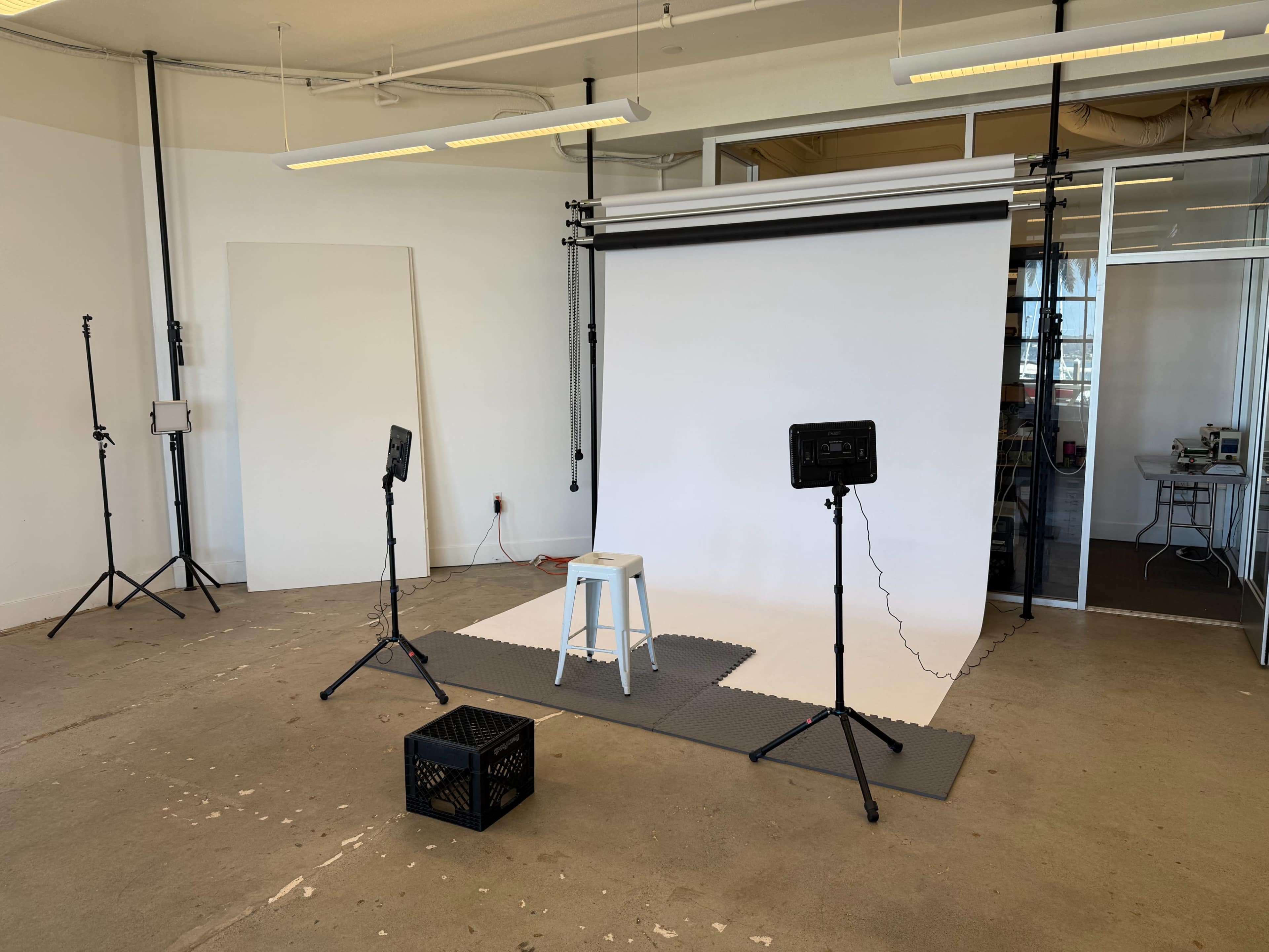 The image shows a photography studio setup with a white backdrop, a stool, lighting equipment on tripods, and a black crate on the floor.