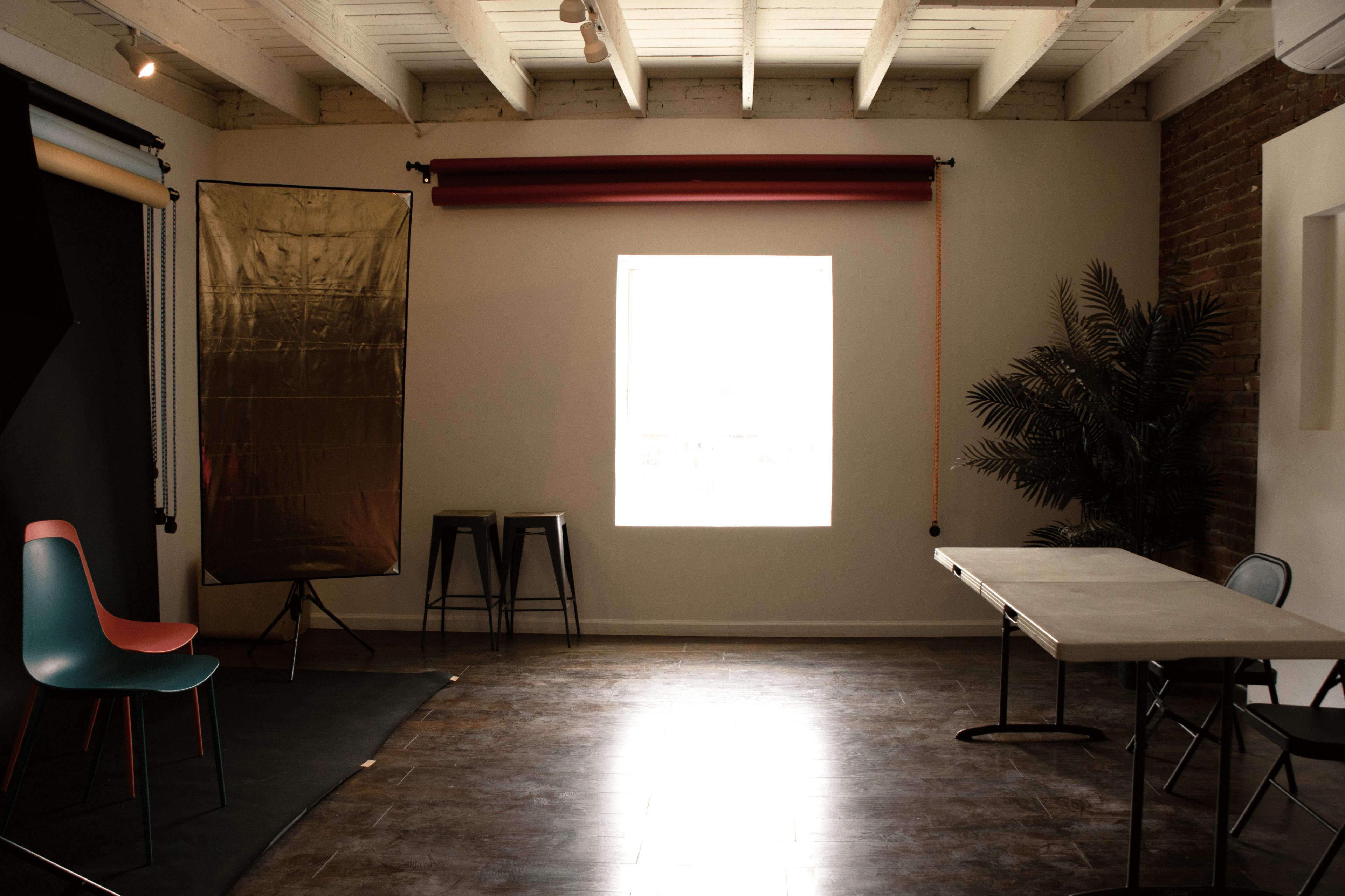A room with wooden floors featuring a table and chairs, a backdrop setup for photography, and a large window providing natural light.