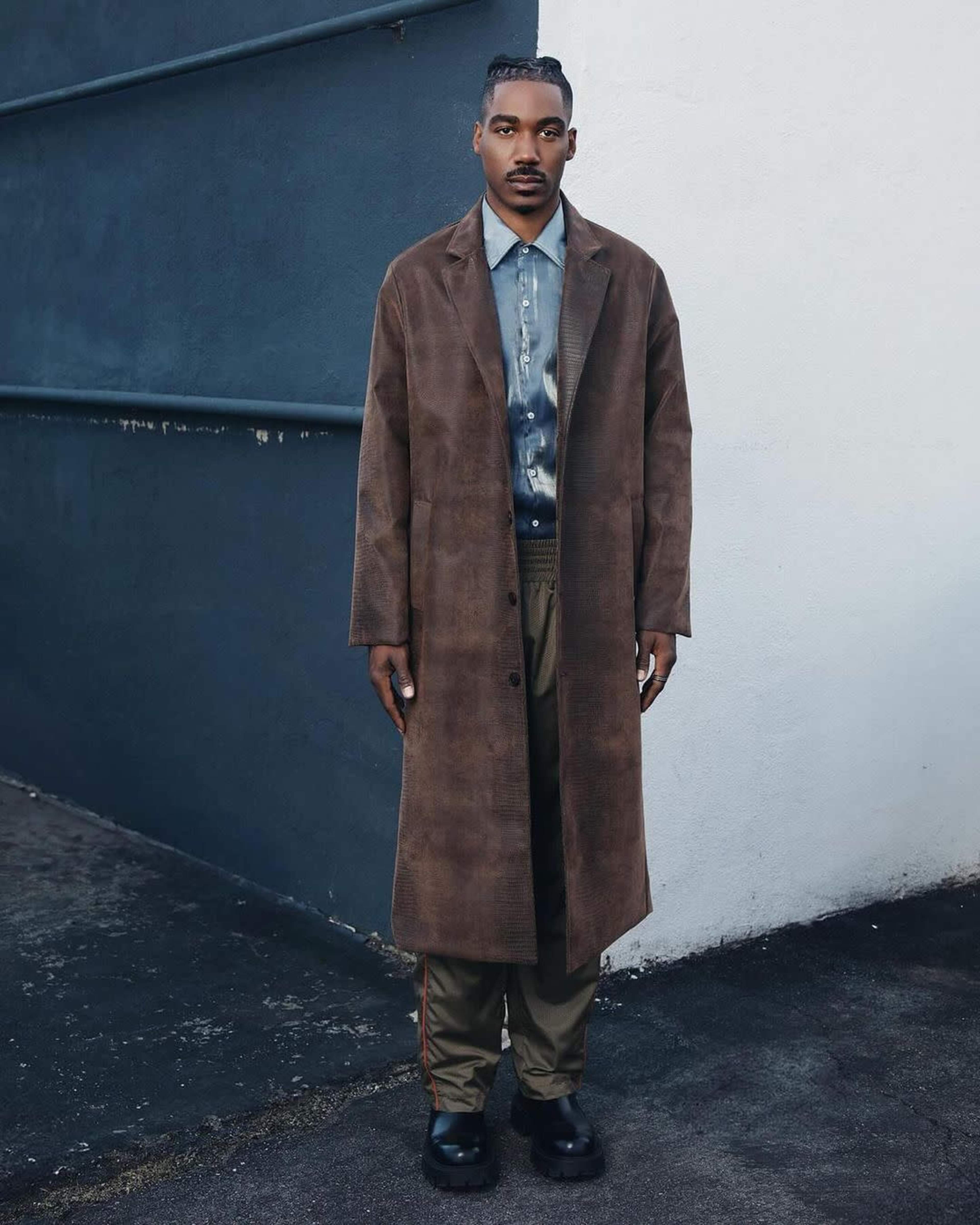 A person stands against a plain wall wearing a long brown coat over a blue shirt and green pants, with black shoes.