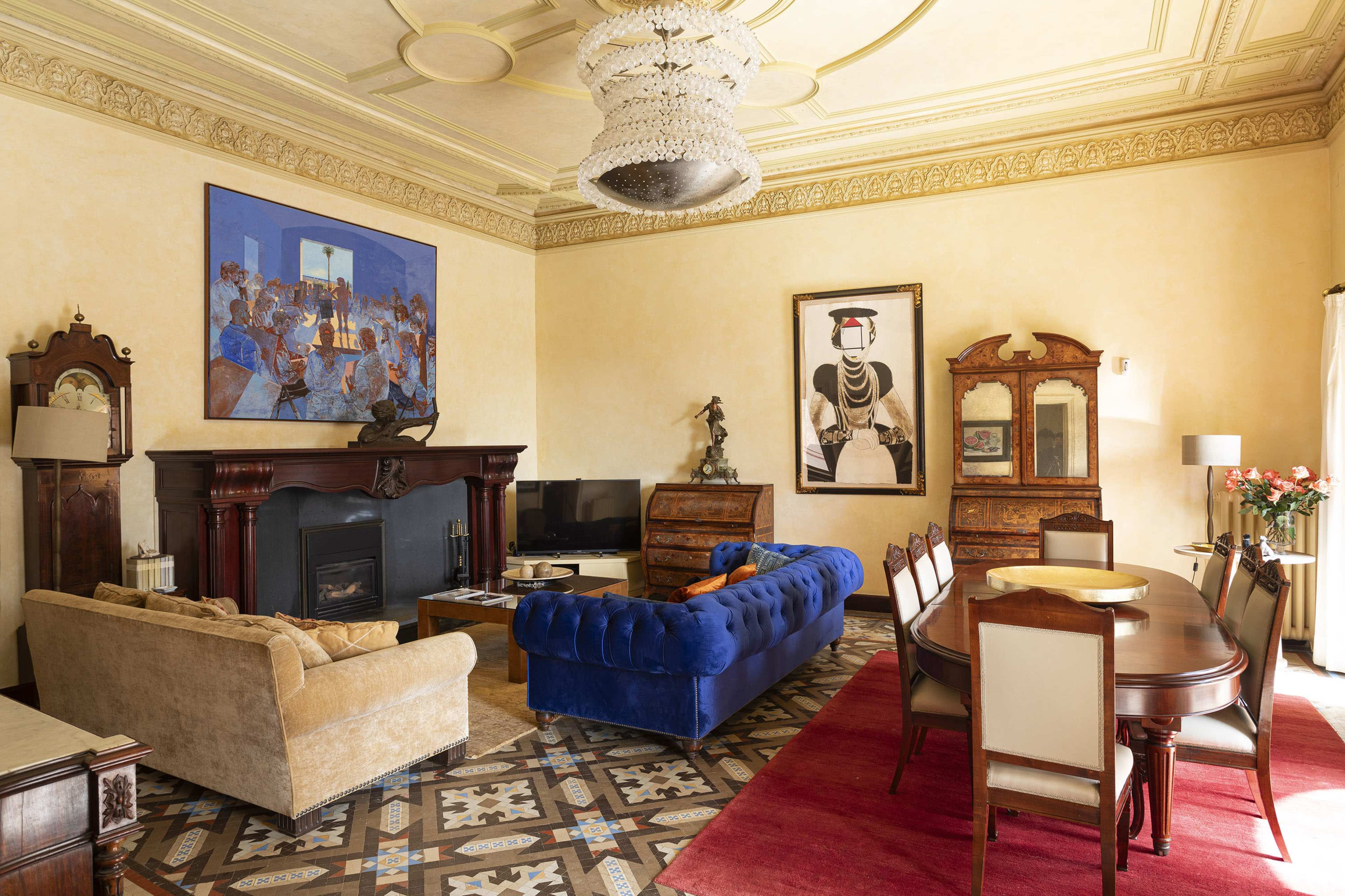 The room features a blue velvet sofa, a beige couch, a wooden dining table, and various artworks on the walls, with a chandelier above and patterned flooring.
