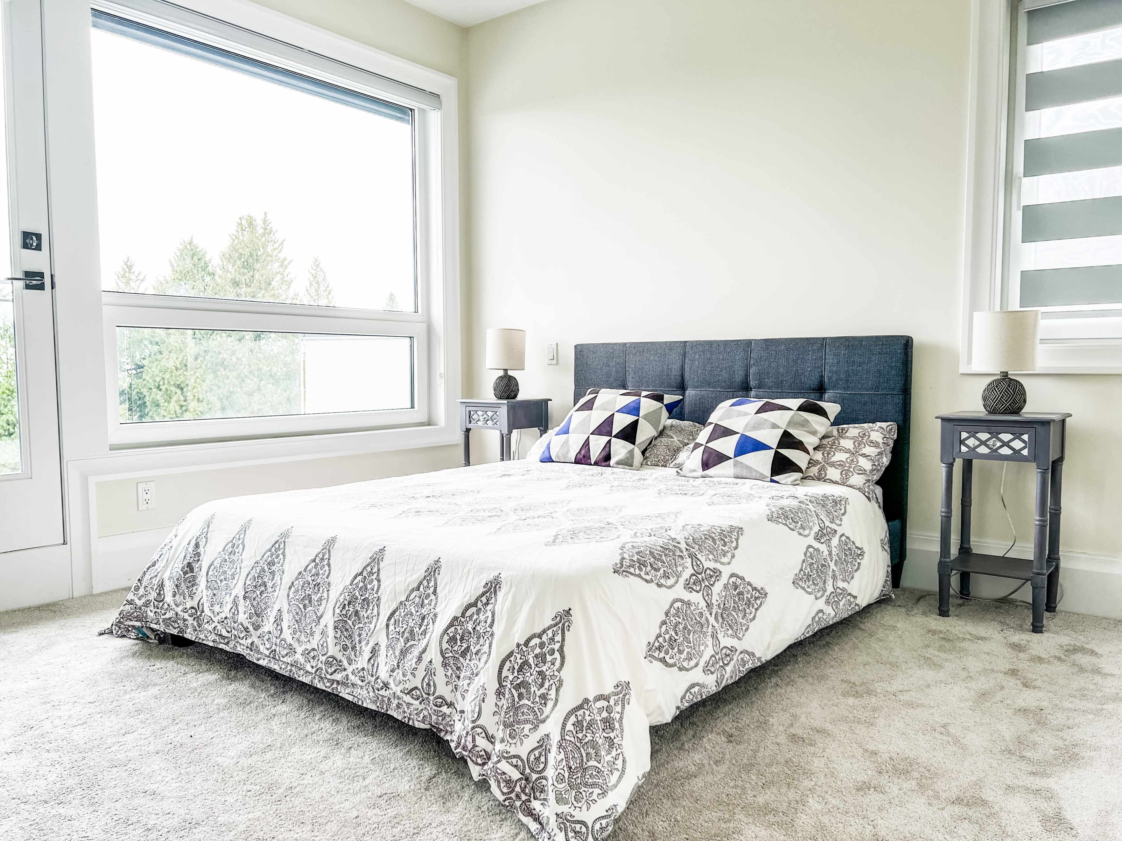 A modern bedroom with a low-profile bed covered in a patterned blanket, flanked by two bedside tables and a large window displaying trees outside.