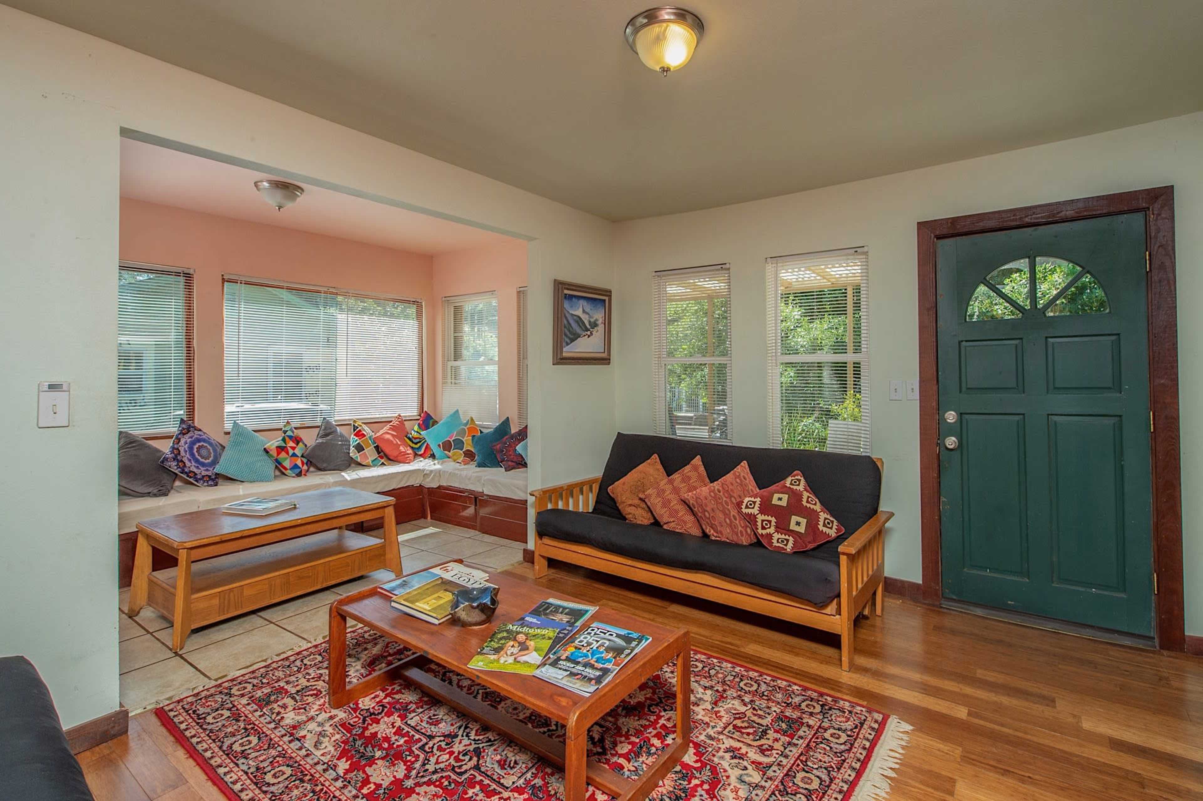 A living room with two sofas, a coffee table, and a window seat adorned with colorful cushions, alongside a green front door.