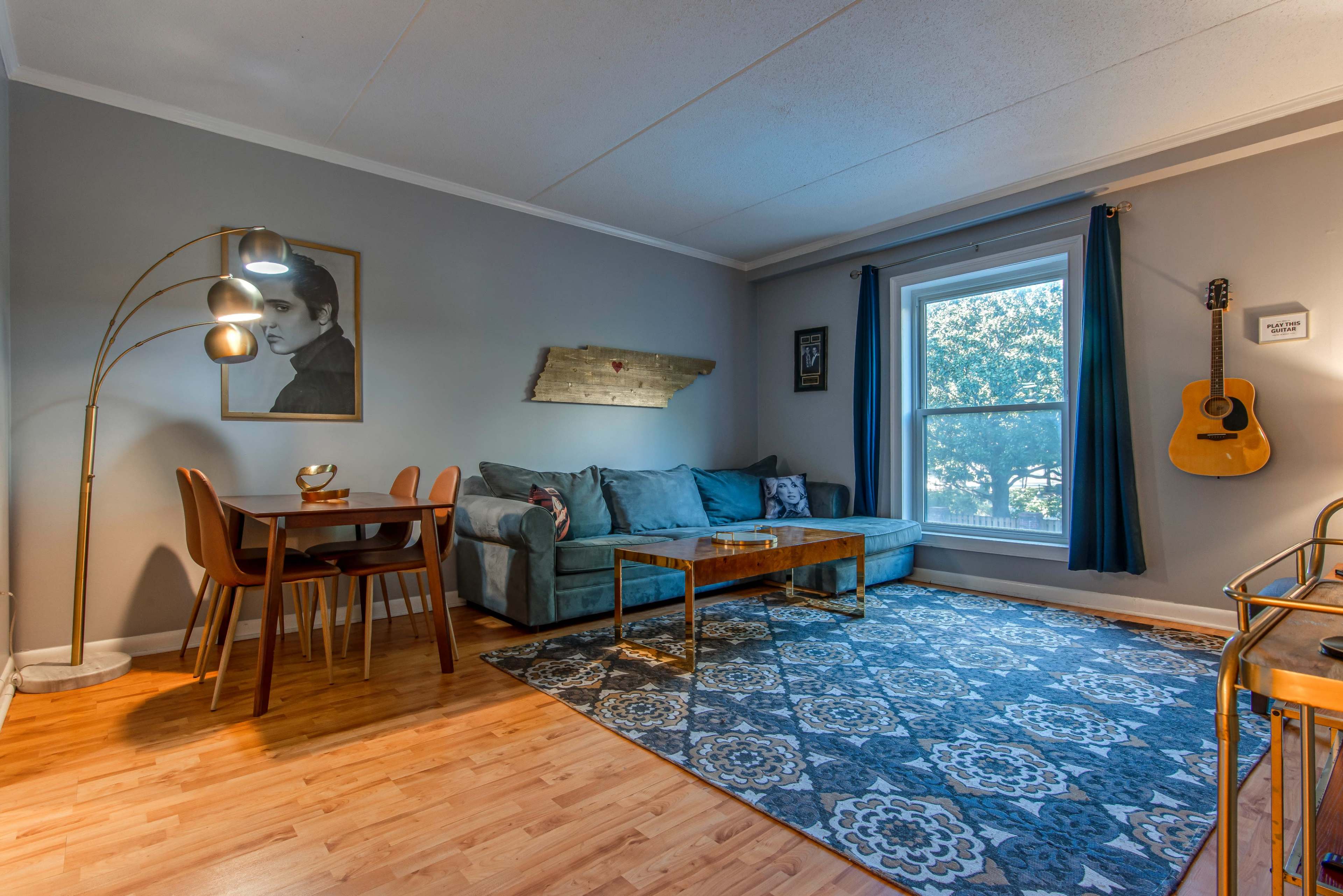 A living room with a teal sofa, a wooden coffee table, a dining area with chairs, and a large window with natural light.