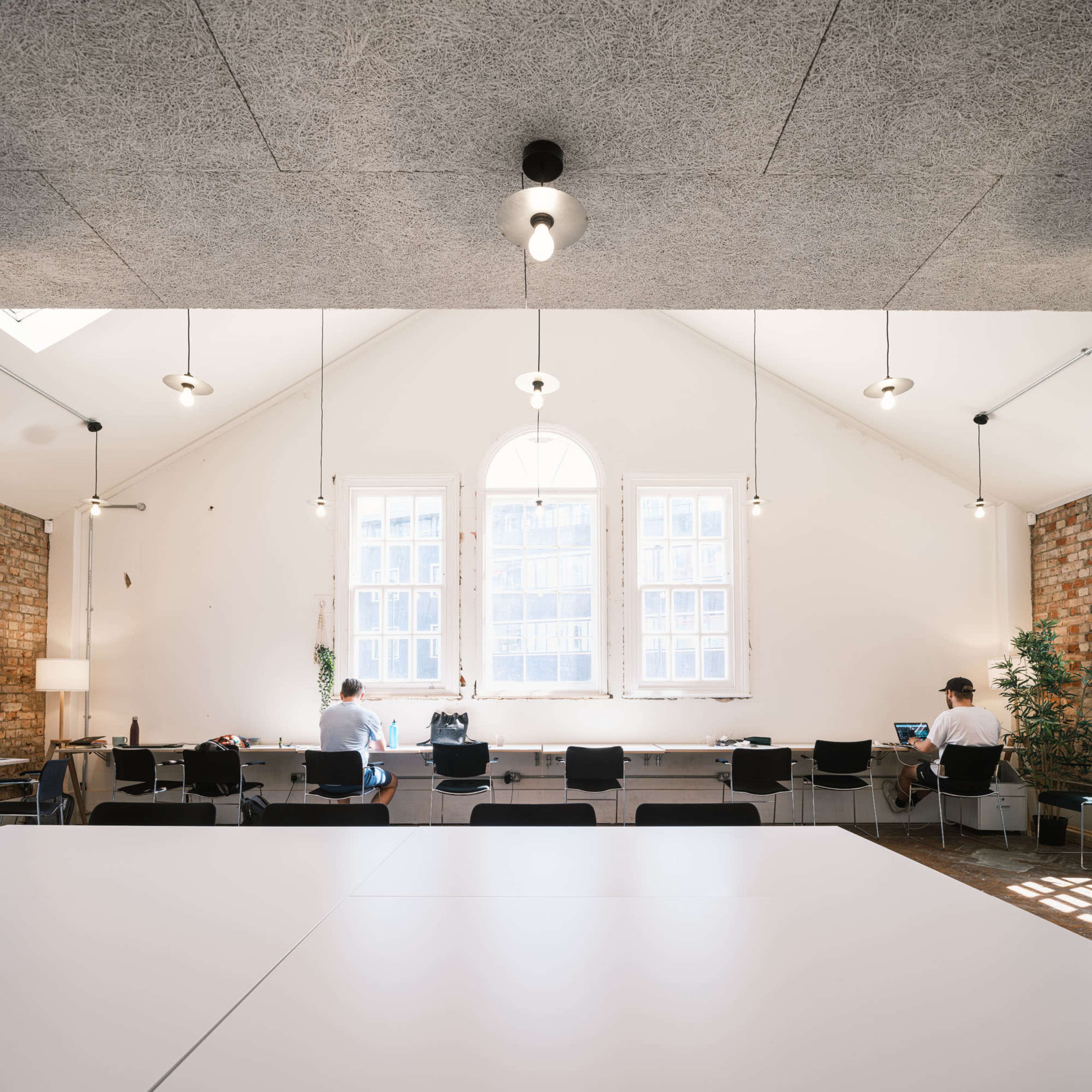A spacious room with a vaulted ceiling, featuring long tables, hanging lights, and two individuals working at laptops near large windows.