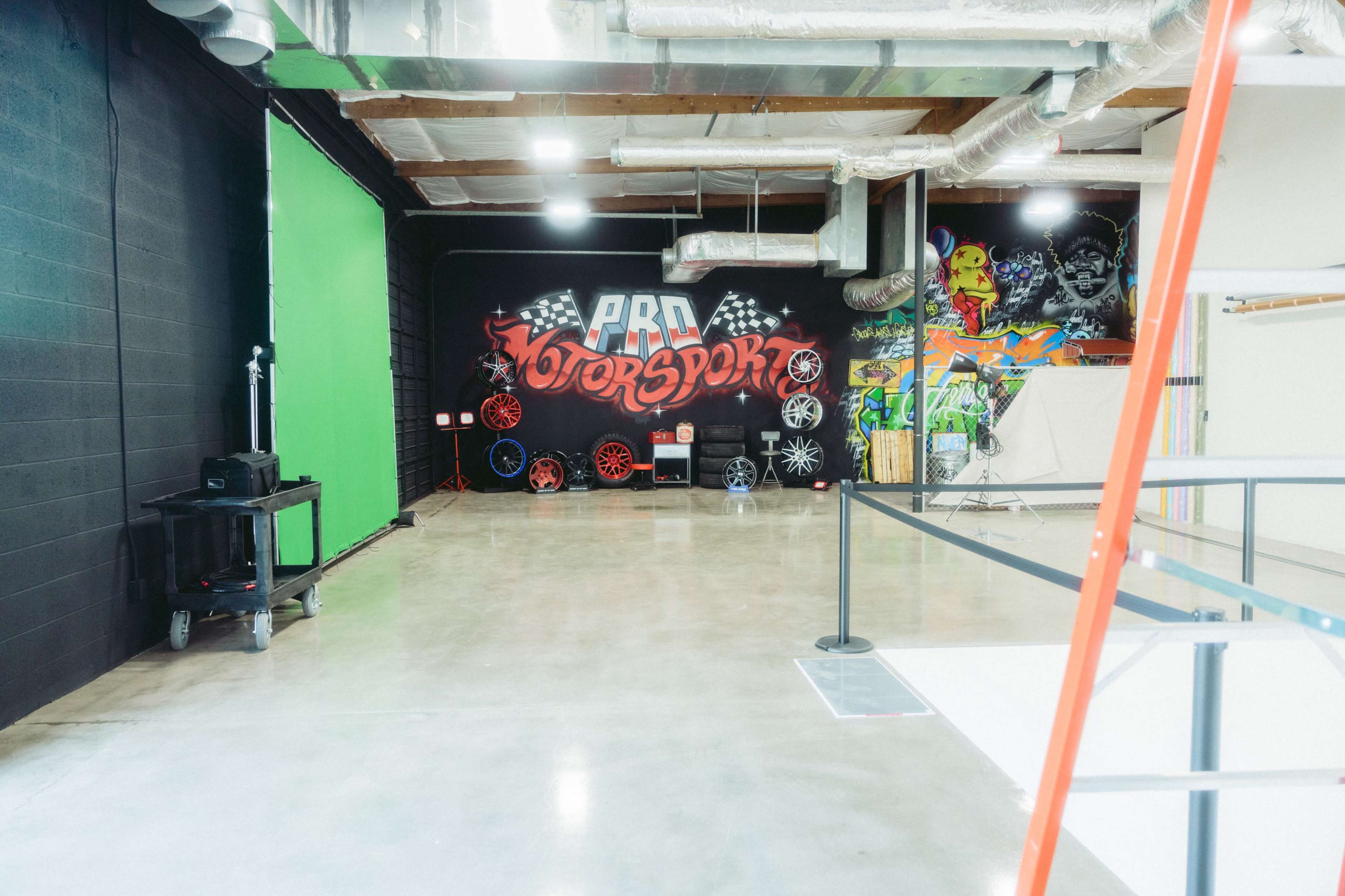 The image shows an indoor space featuring a colorful mural with automotive themes, a green screen area, and a collection of wheels against a black wall.