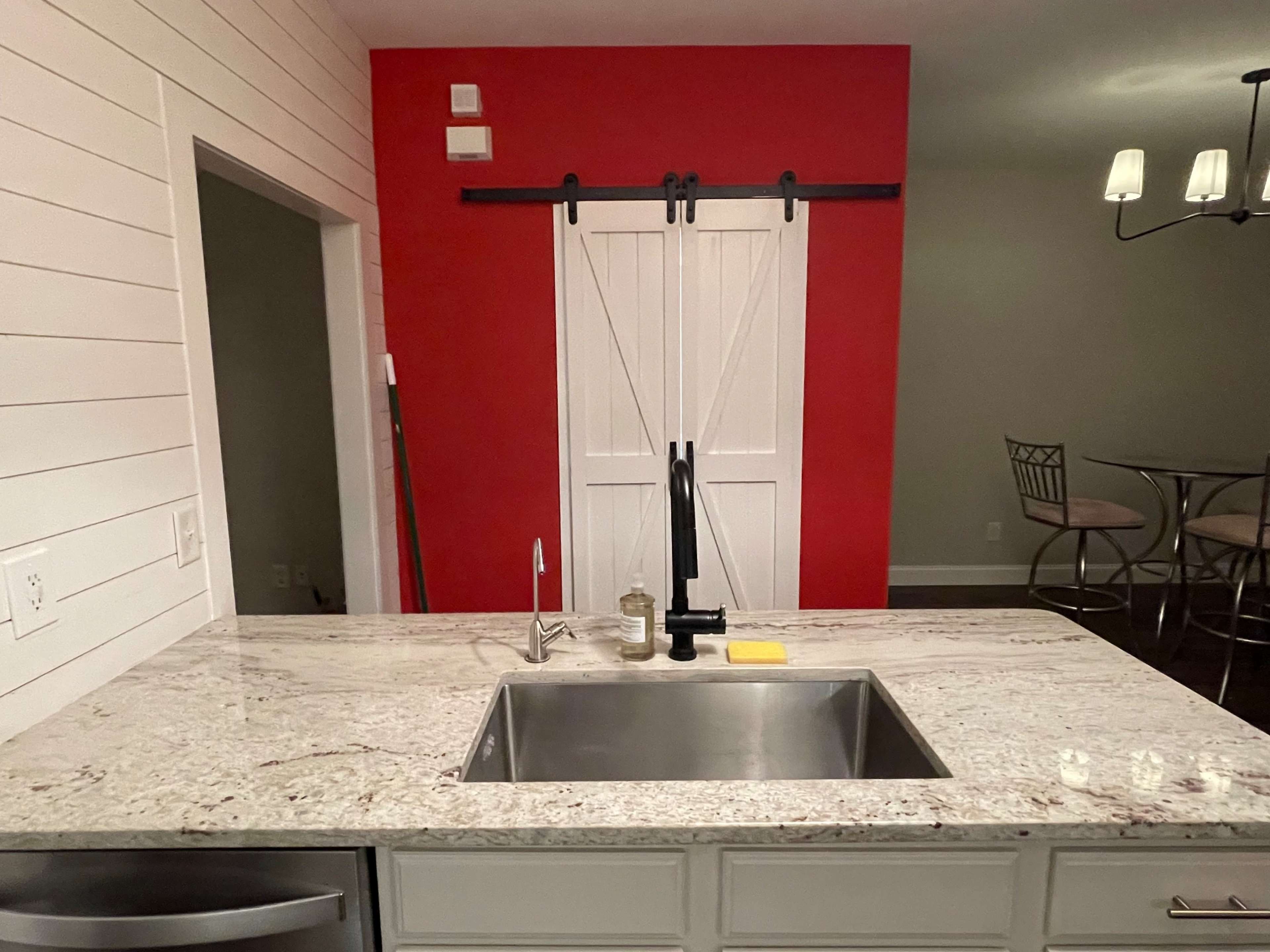 A modern kitchen features a stainless steel sink set in a light-colored granite countertop, with a red accent wall and a sliding barn door in the background.