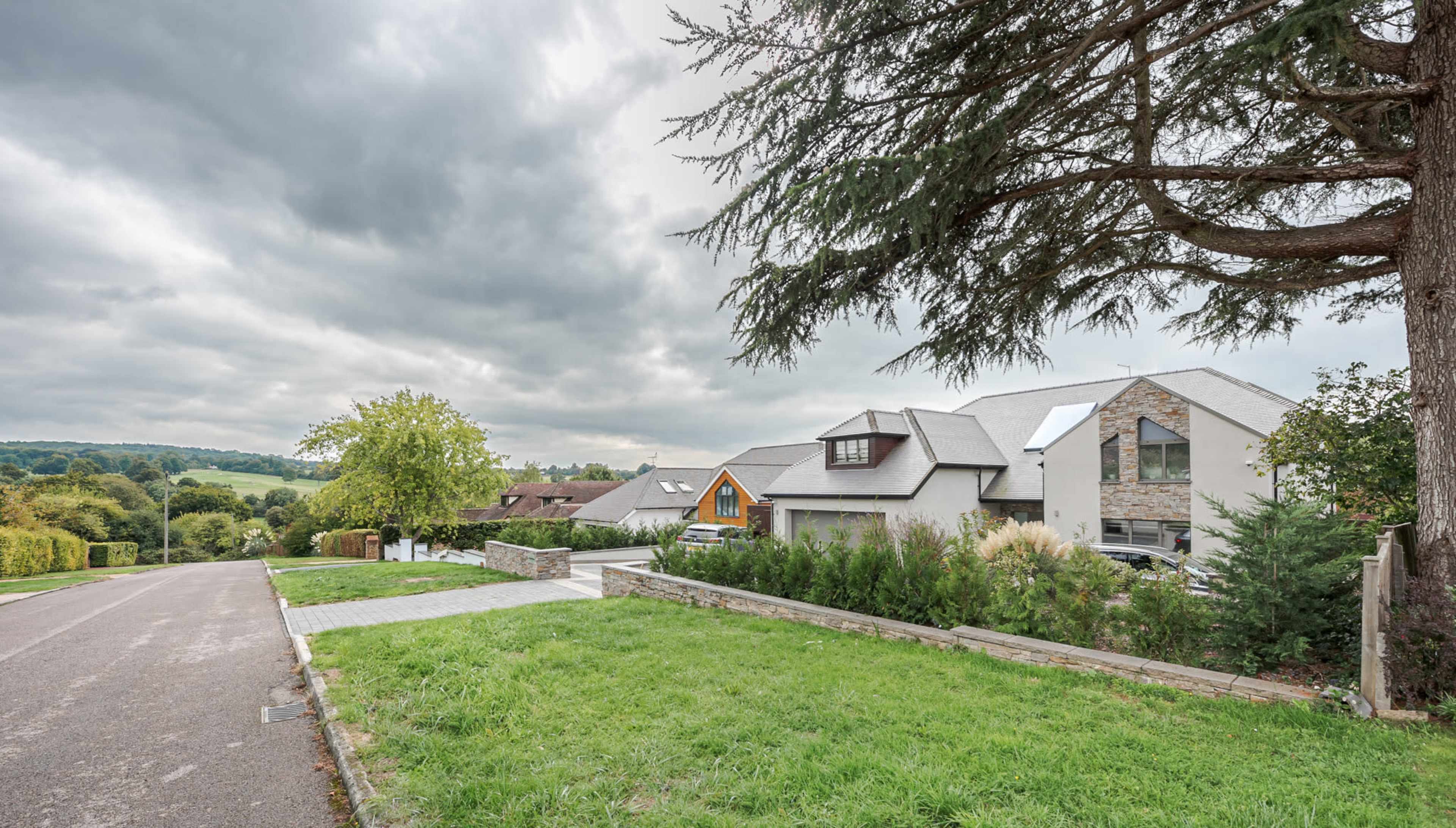A modern house with a stone facade is situated alongside a tree-lined road, overlooking a green landscape under a cloudy sky.