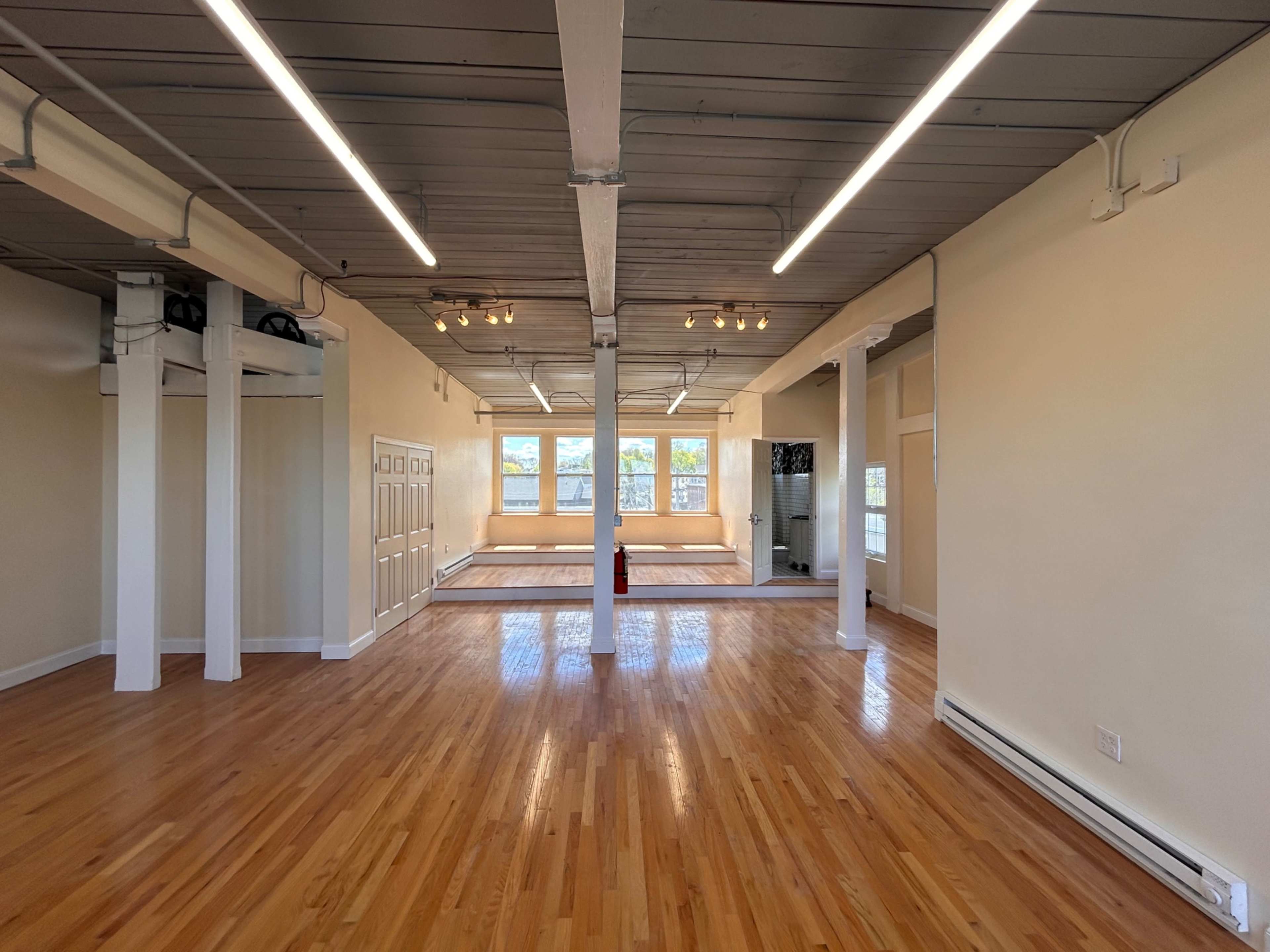 The image shows an empty room with polished wooden floors, large windows letting in natural light, and exposed wooden beams on the ceiling.