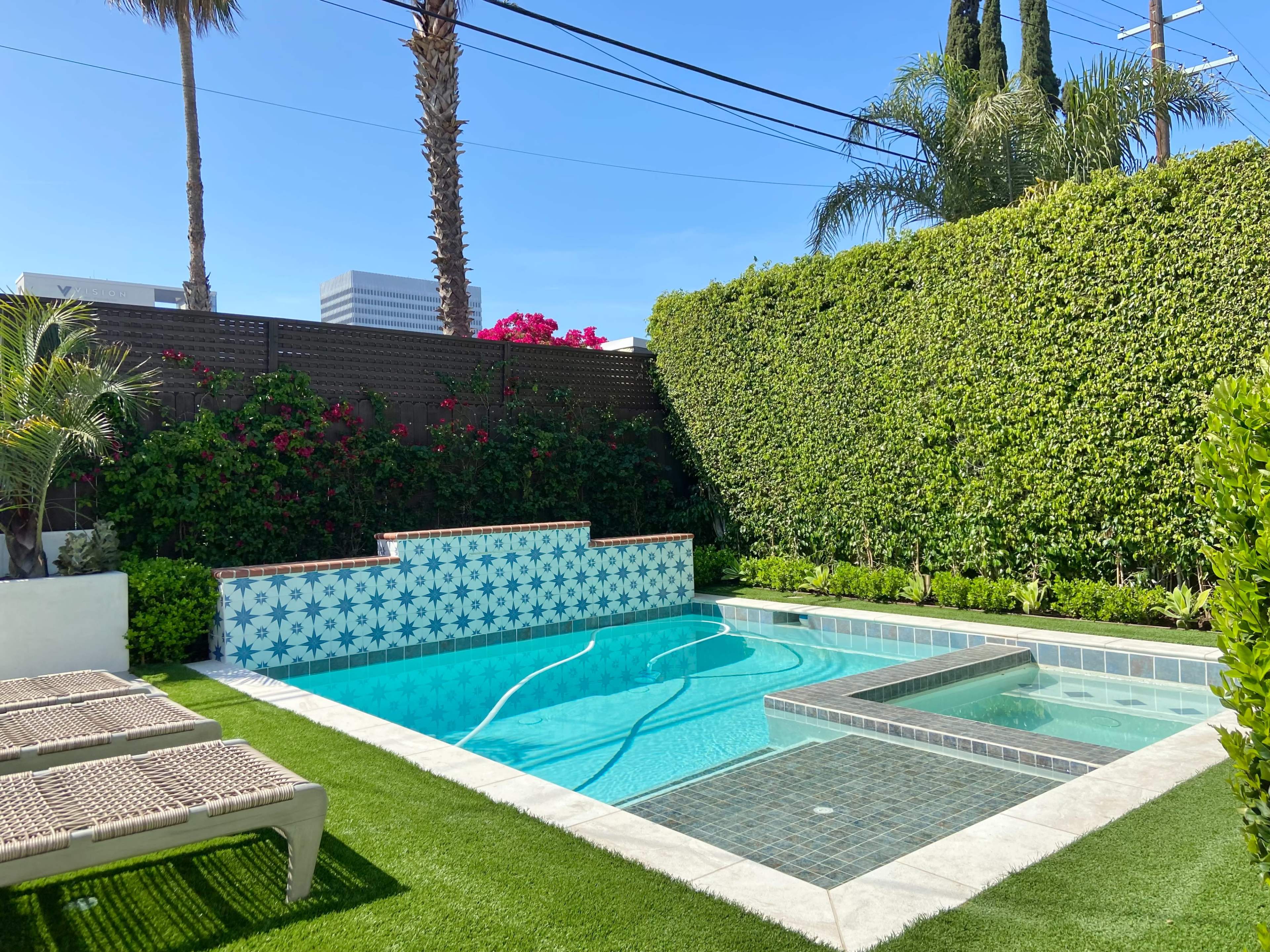 The image shows a backyard pool area featuring a tiled swimming pool, a spa, and landscaped greenery surrounded by palm trees and flowering plants.