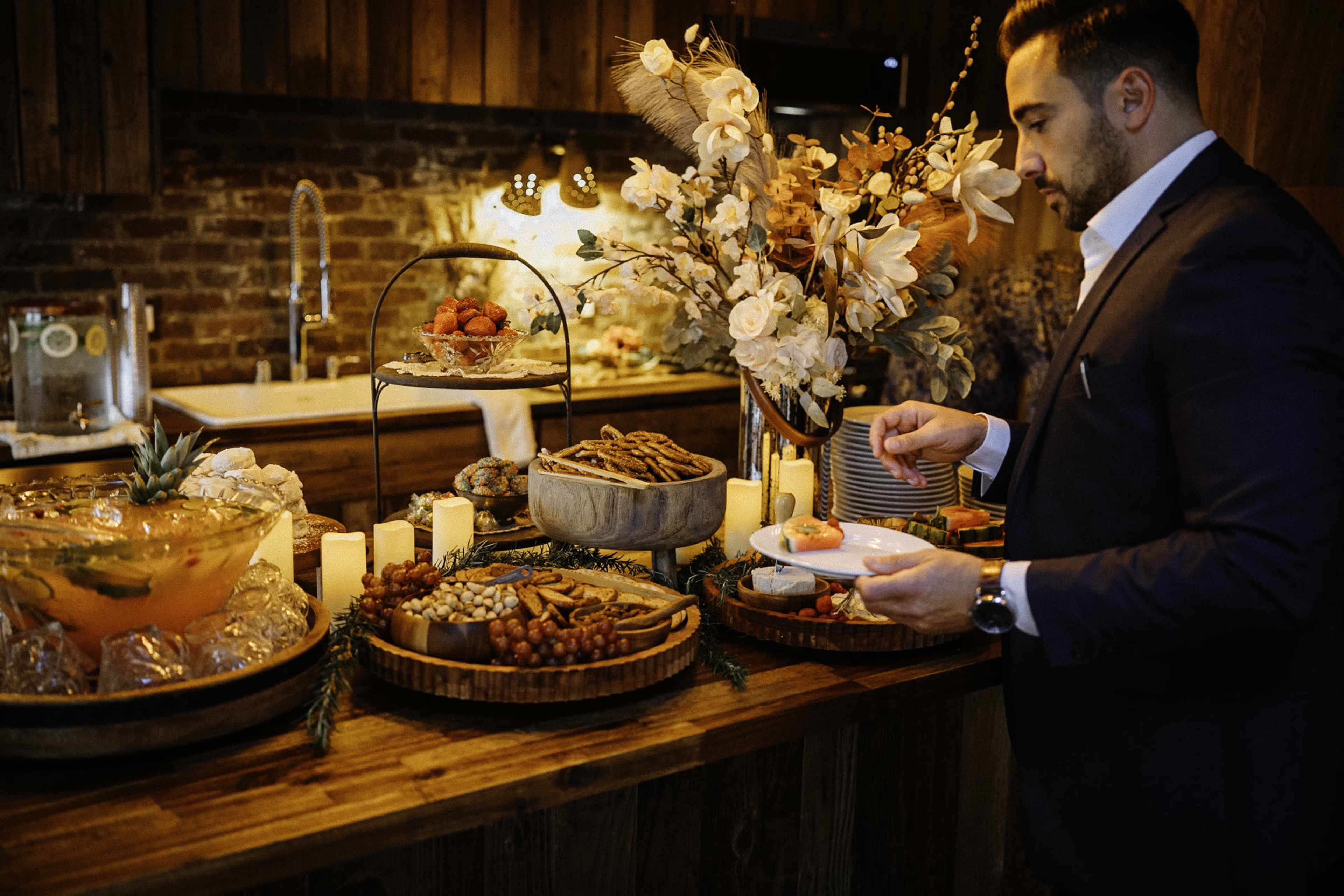 A man is serving himself food from a buffet table decorated with various appetizers and floral arrangements in a warmly lit room.