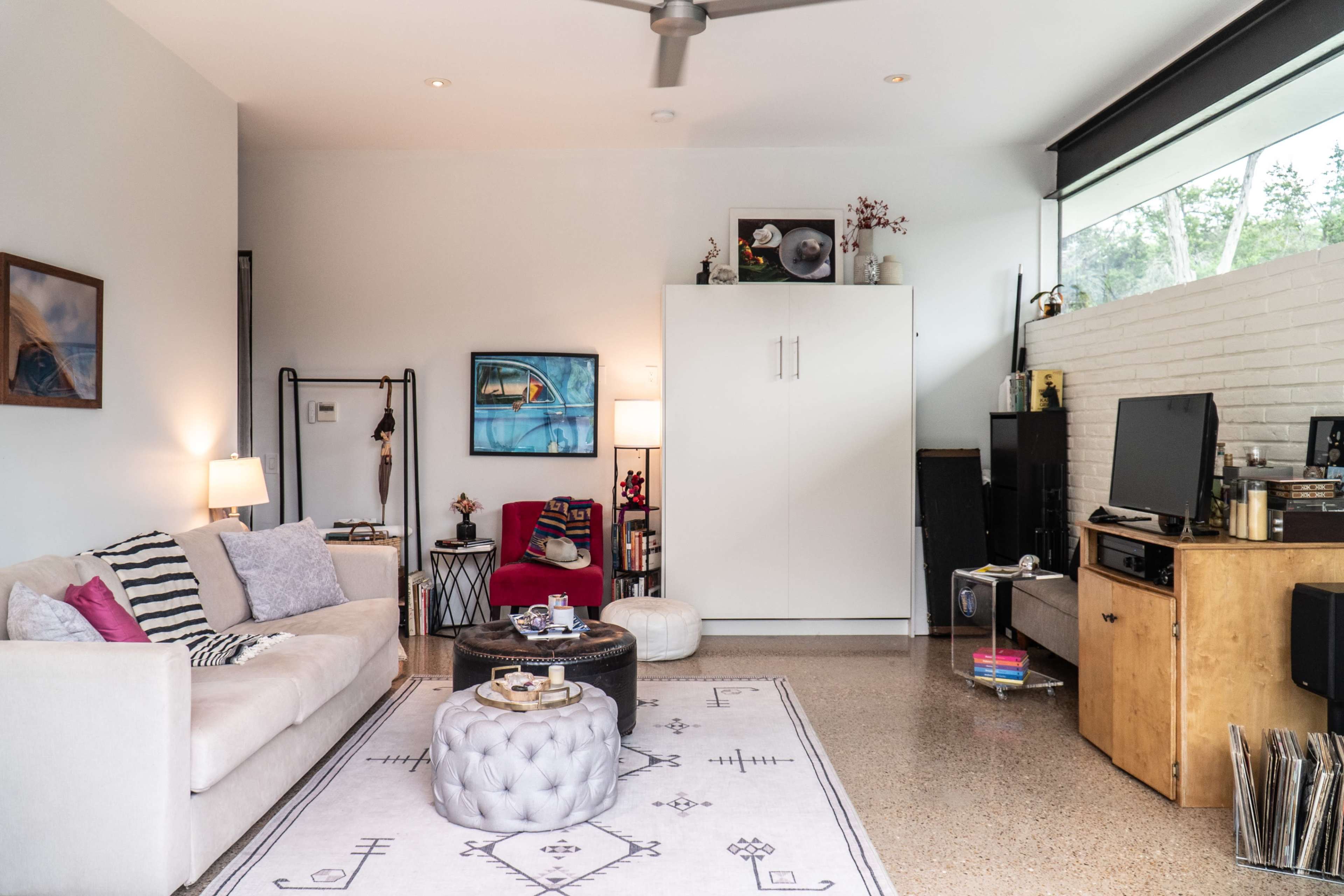 A light-filled living room features a beige sofa, a coffee table, and various shelves with decorations and a television against a white brick wall.
