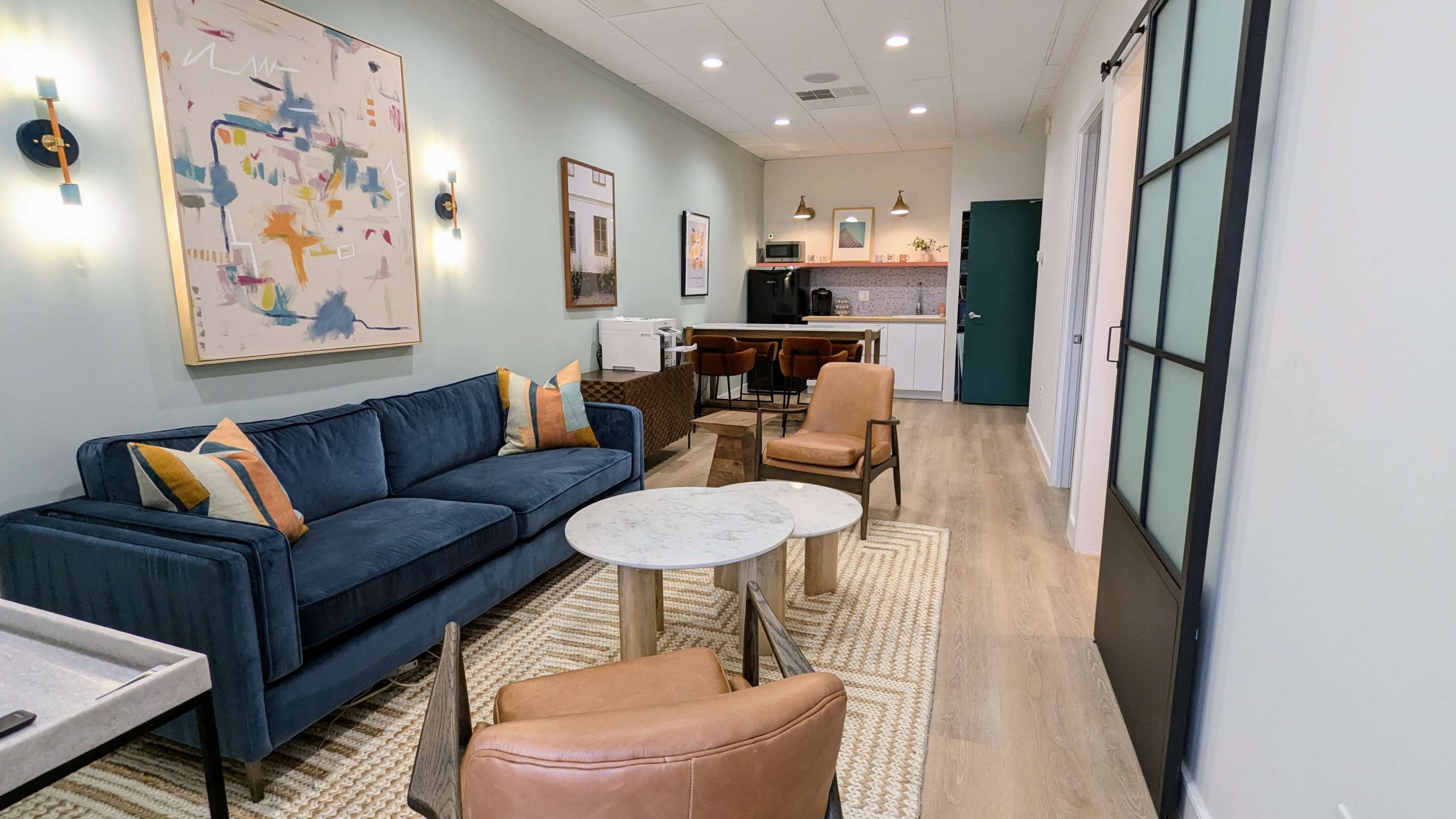 A well-lit, modern living space featuring a blue sofa, brown armchairs, a marble coffee table, and a kitchenette in the background.