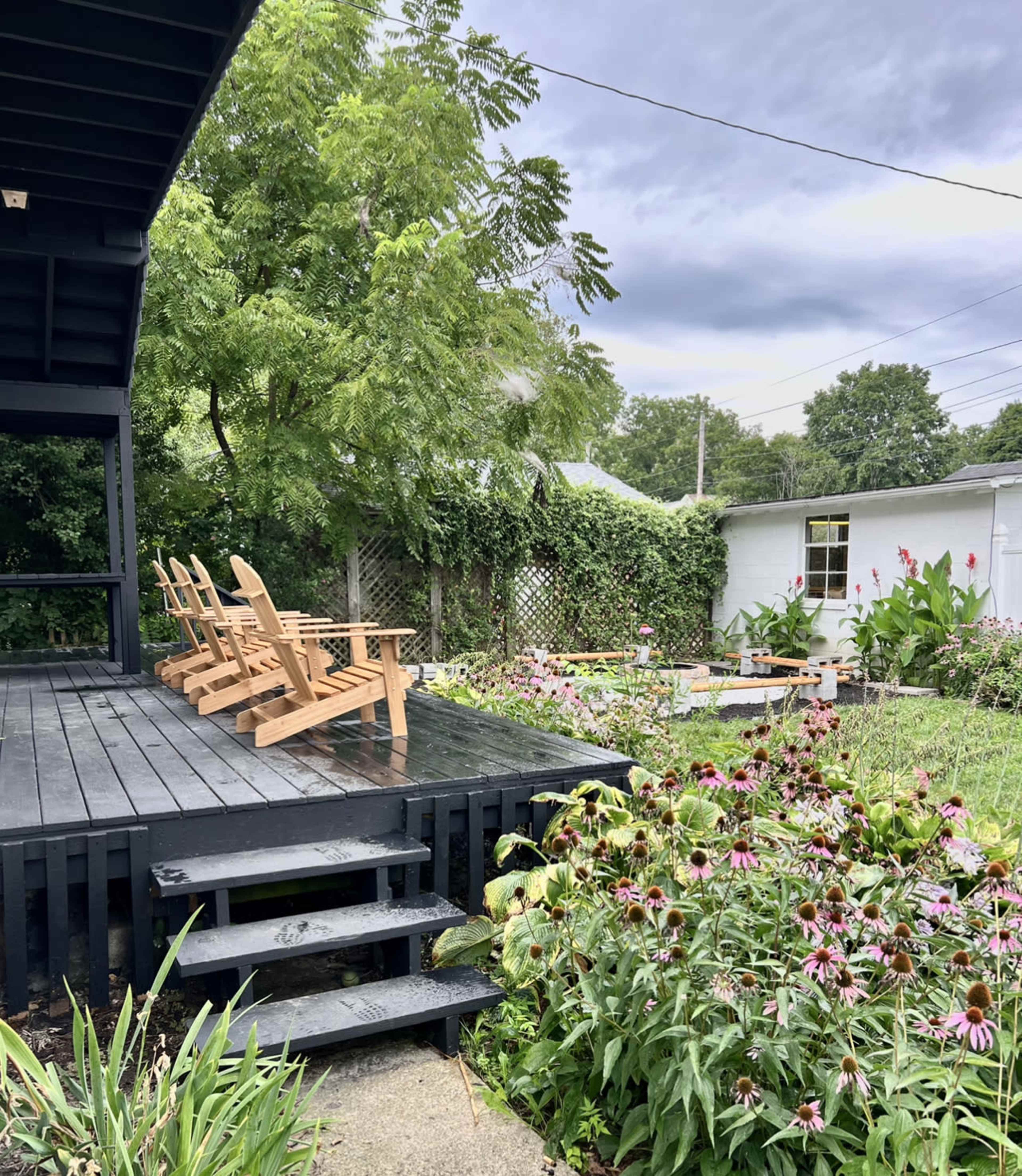 A wooden deck with several Adirondack chairs overlooks a garden filled with blooming flowers and greenery.