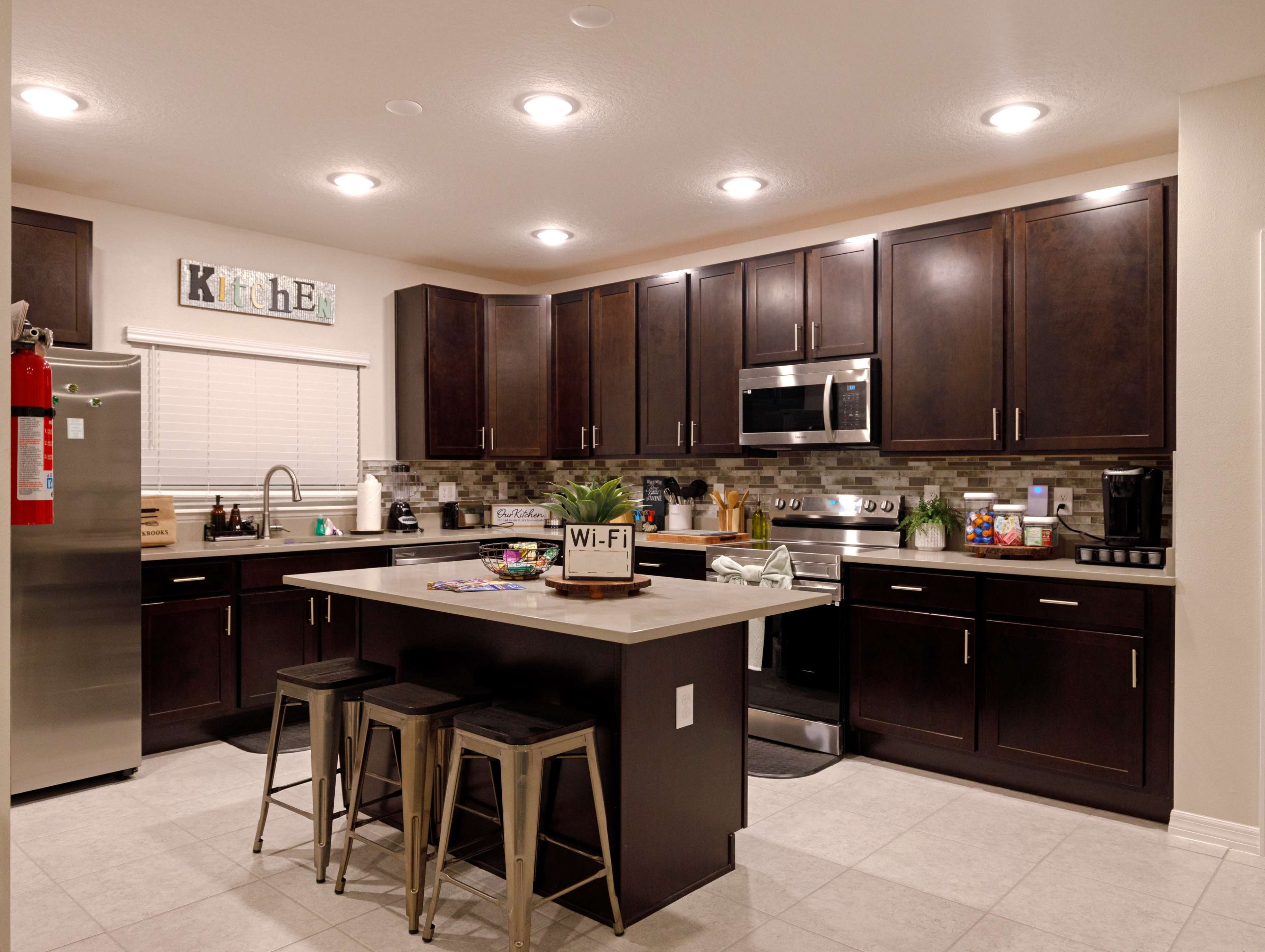 A modern kitchen with dark wood cabinets, stainless steel appliances, and a central island with three metal bar stools.