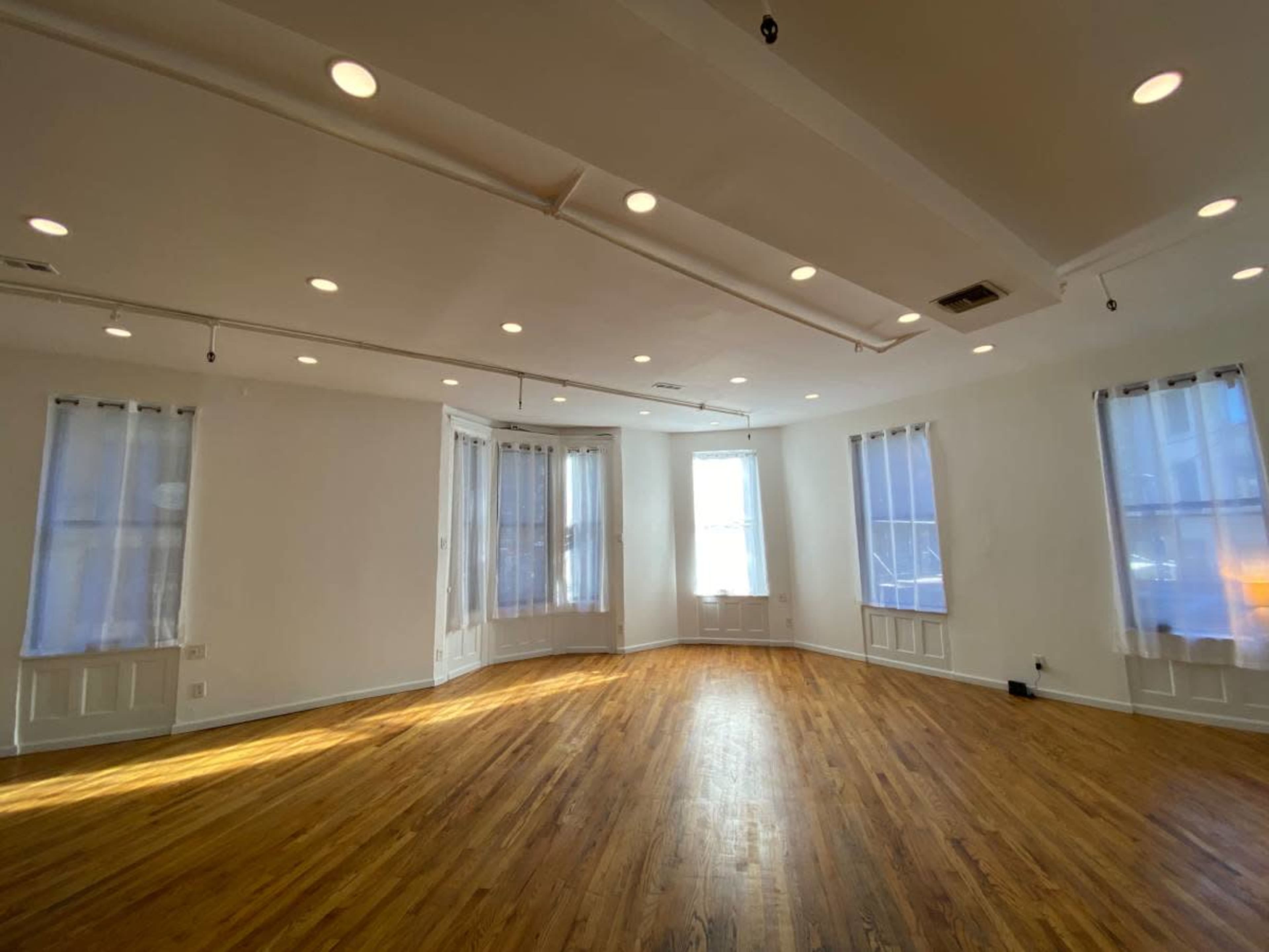 The image shows a spacious, well-lit room with wooden floors, featuring multiple windows and white walls.