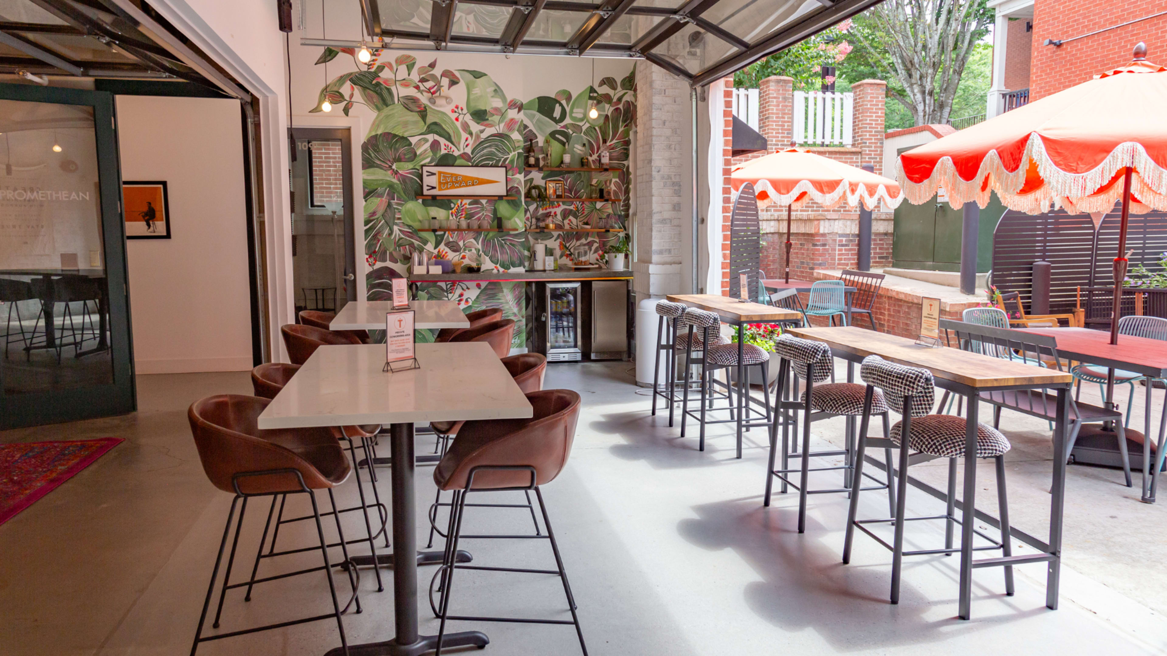 The image shows a modern indoor-outdoor bar area with a tropical-themed mural, seating at high tables, and a large open garage door leading to an outdoor patio.