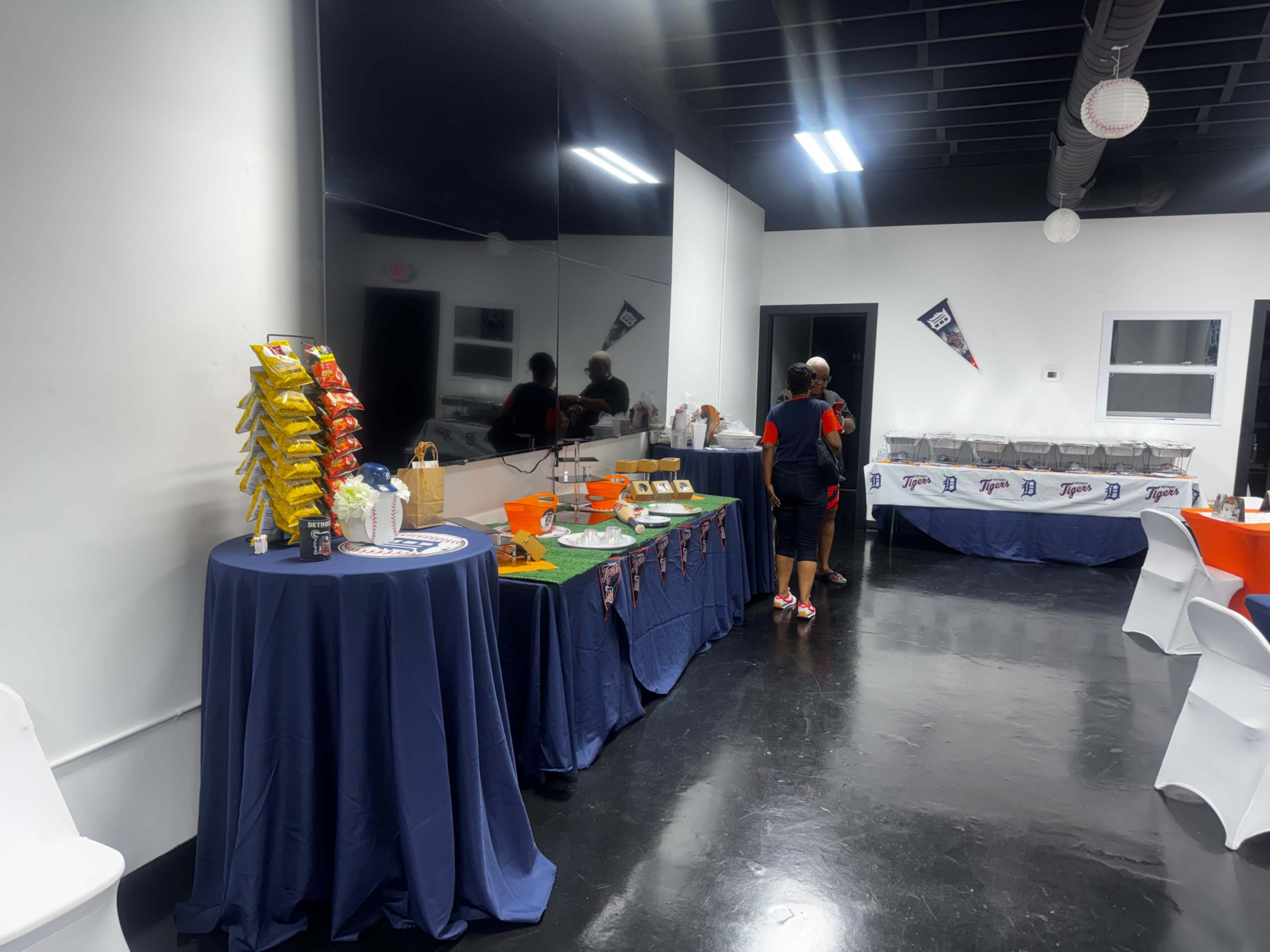 A brightly lit room features a snack table with bags of chips and party supplies, while a person walks towards a serving area with covered dishes.