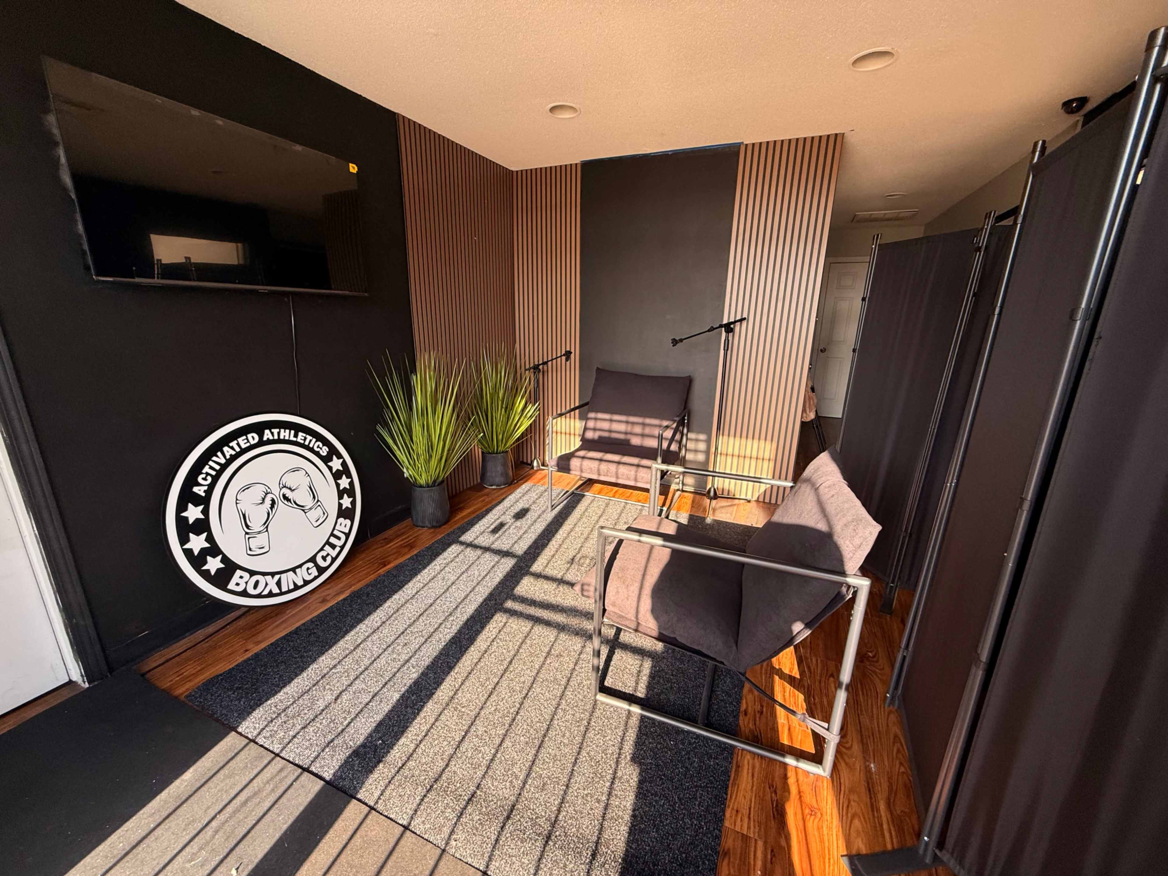 The image shows an interior space of a boxing gym featuring two chairs, a large television, and decorative plants against a backdrop of black and wooden paneling.