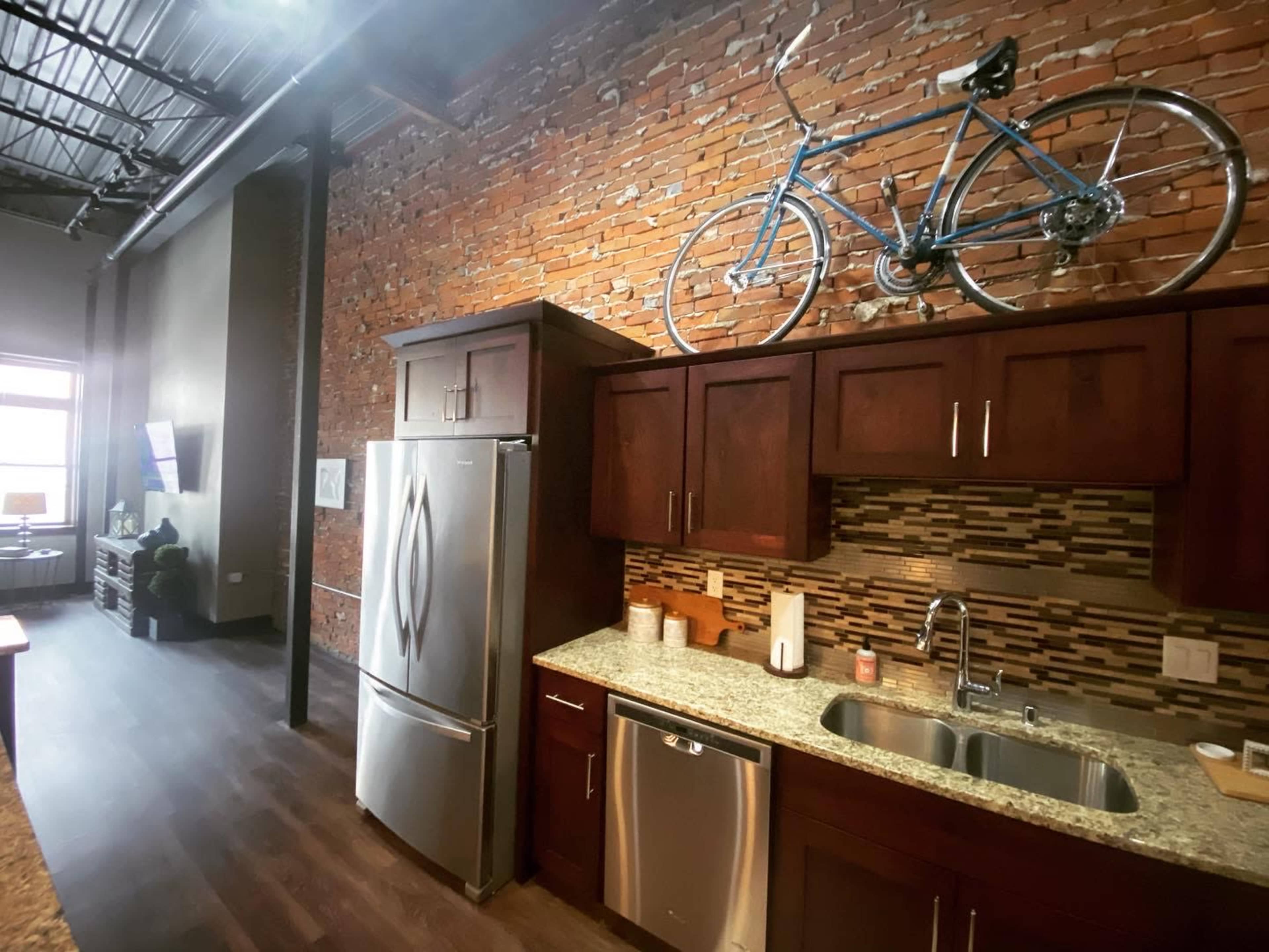 A modern kitchen with wood cabinets, a granite countertop, and a bicycle mounted on a brick wall.