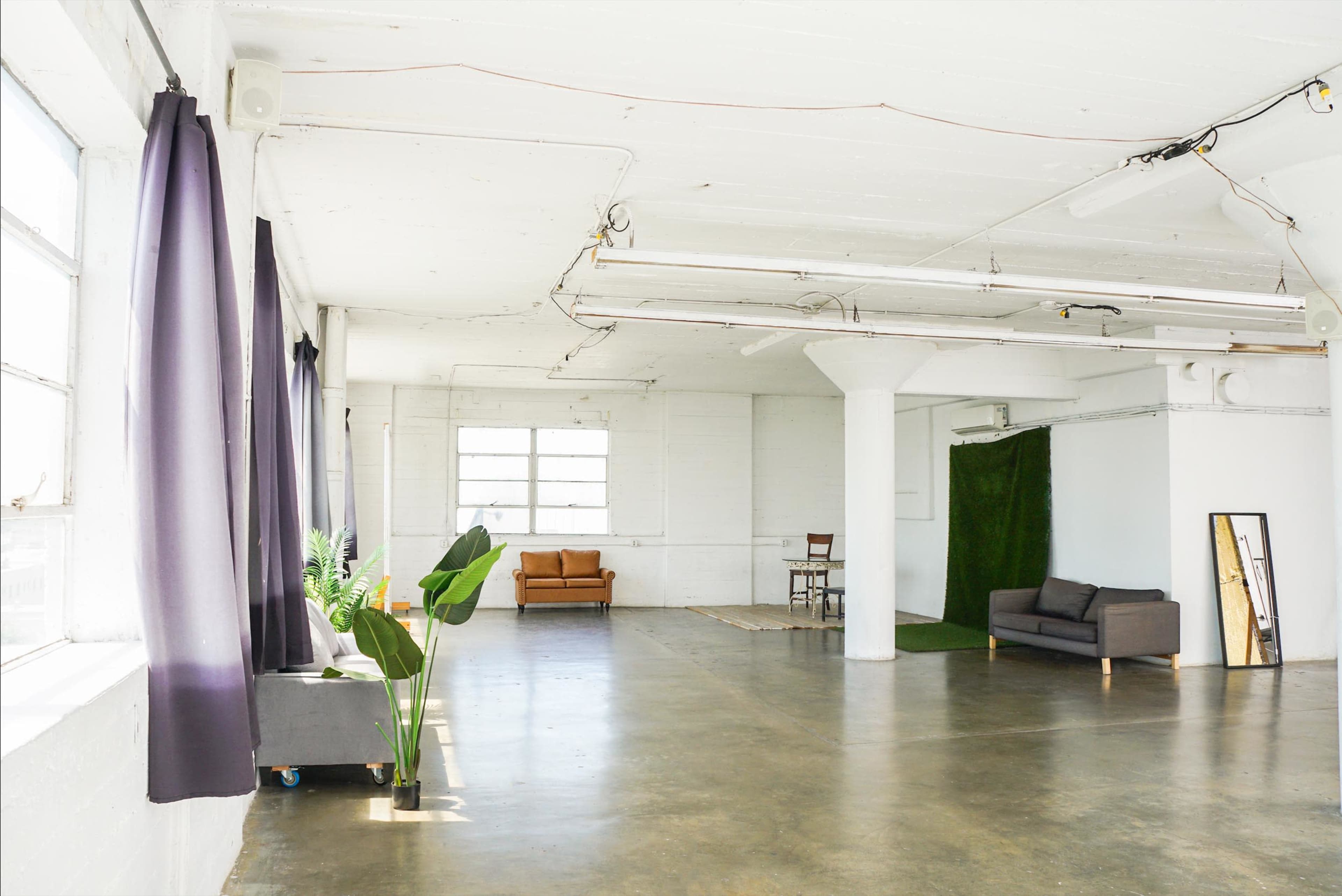 The image shows a spacious, well-lit room with polished concrete floors, featuring a sofa, a rug, and green plants, along with large windows and draped curtains.