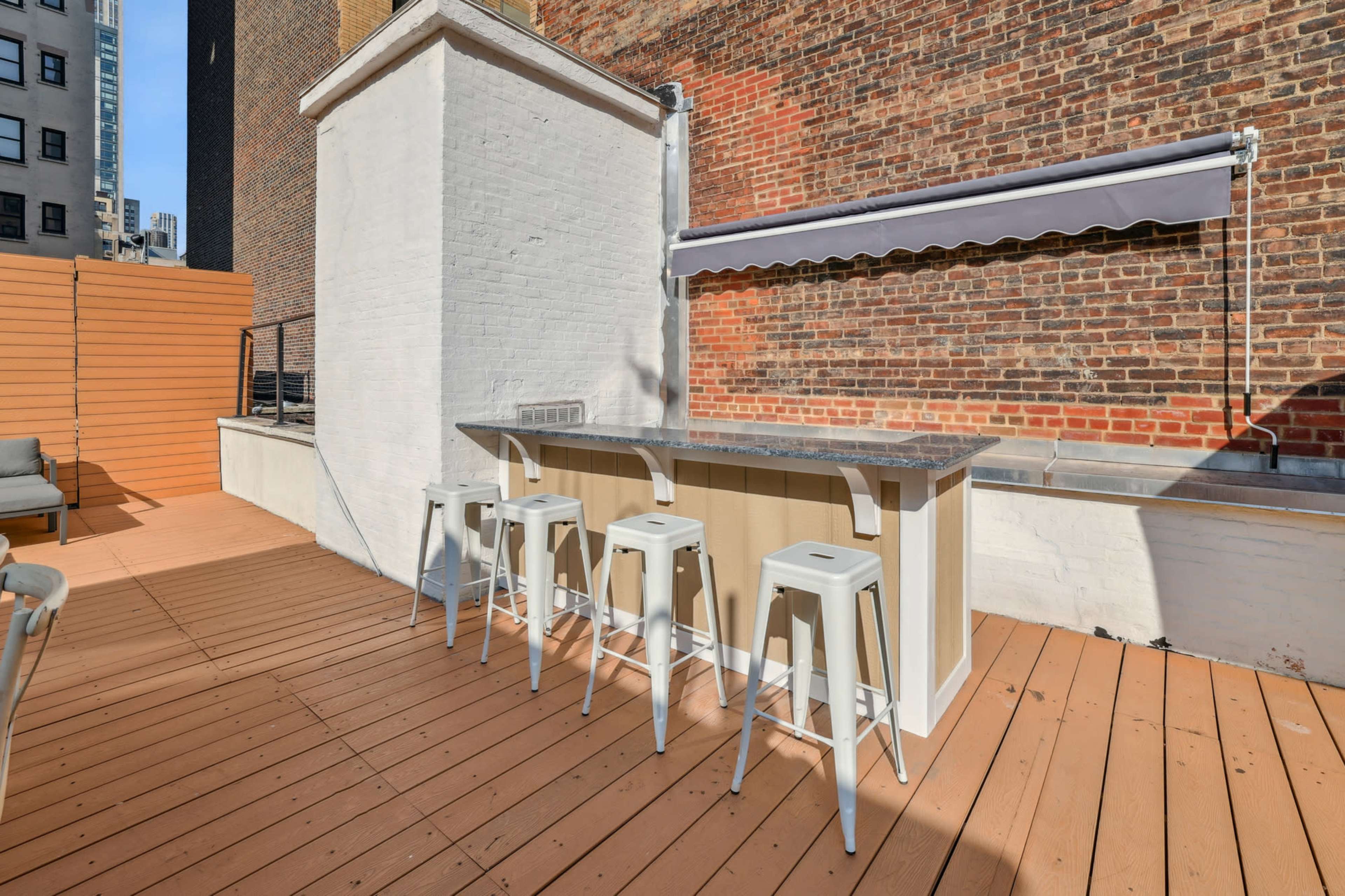 The image shows a rooftop terrace with a wooden deck, featuring a bar counter with three white stools and a retractable awning.