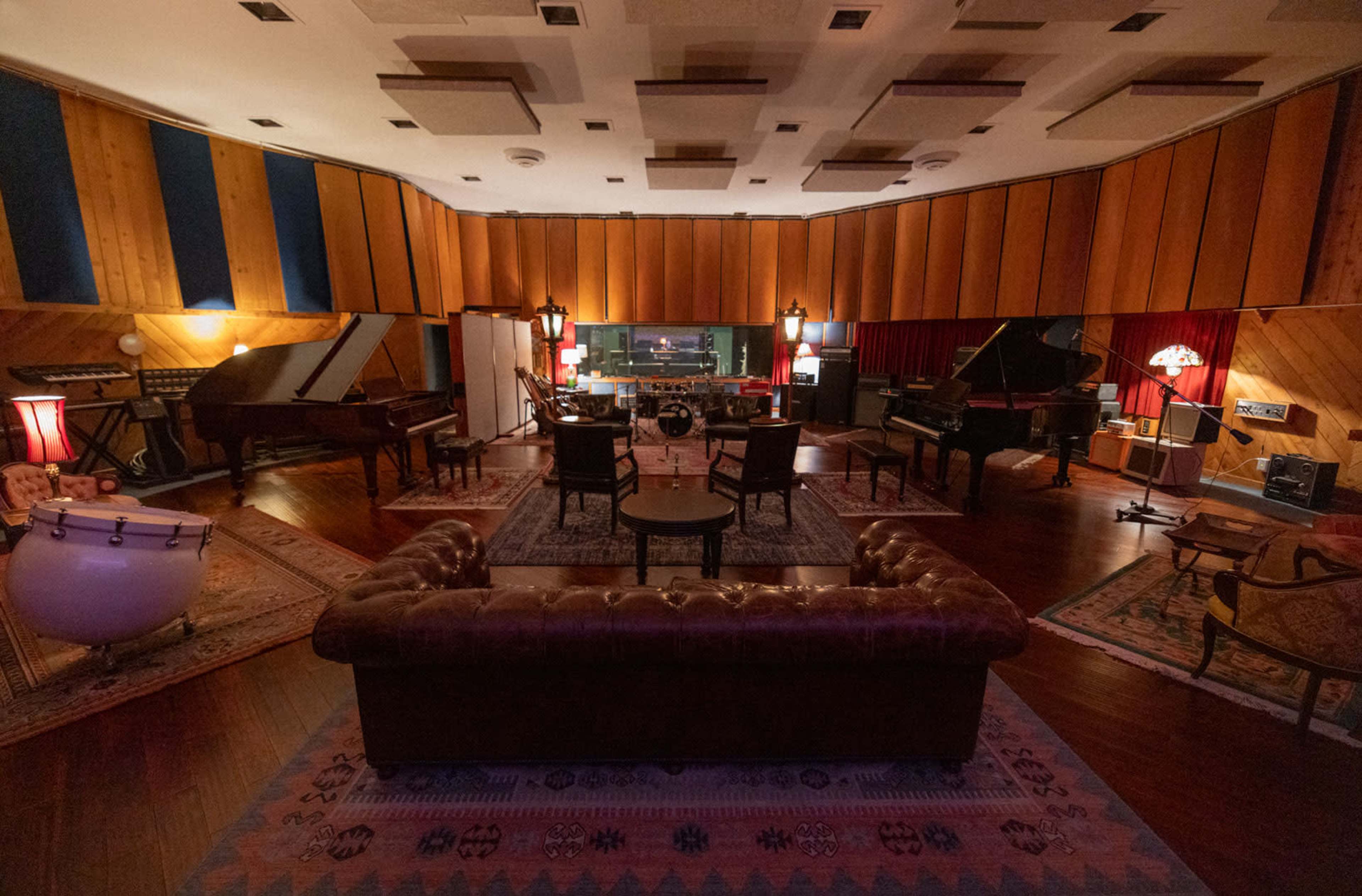 The image shows a vintage recording studio with multiple grand pianos, a mixing console in the background, and a cozy seating area featuring a leather sofa and several chairs.