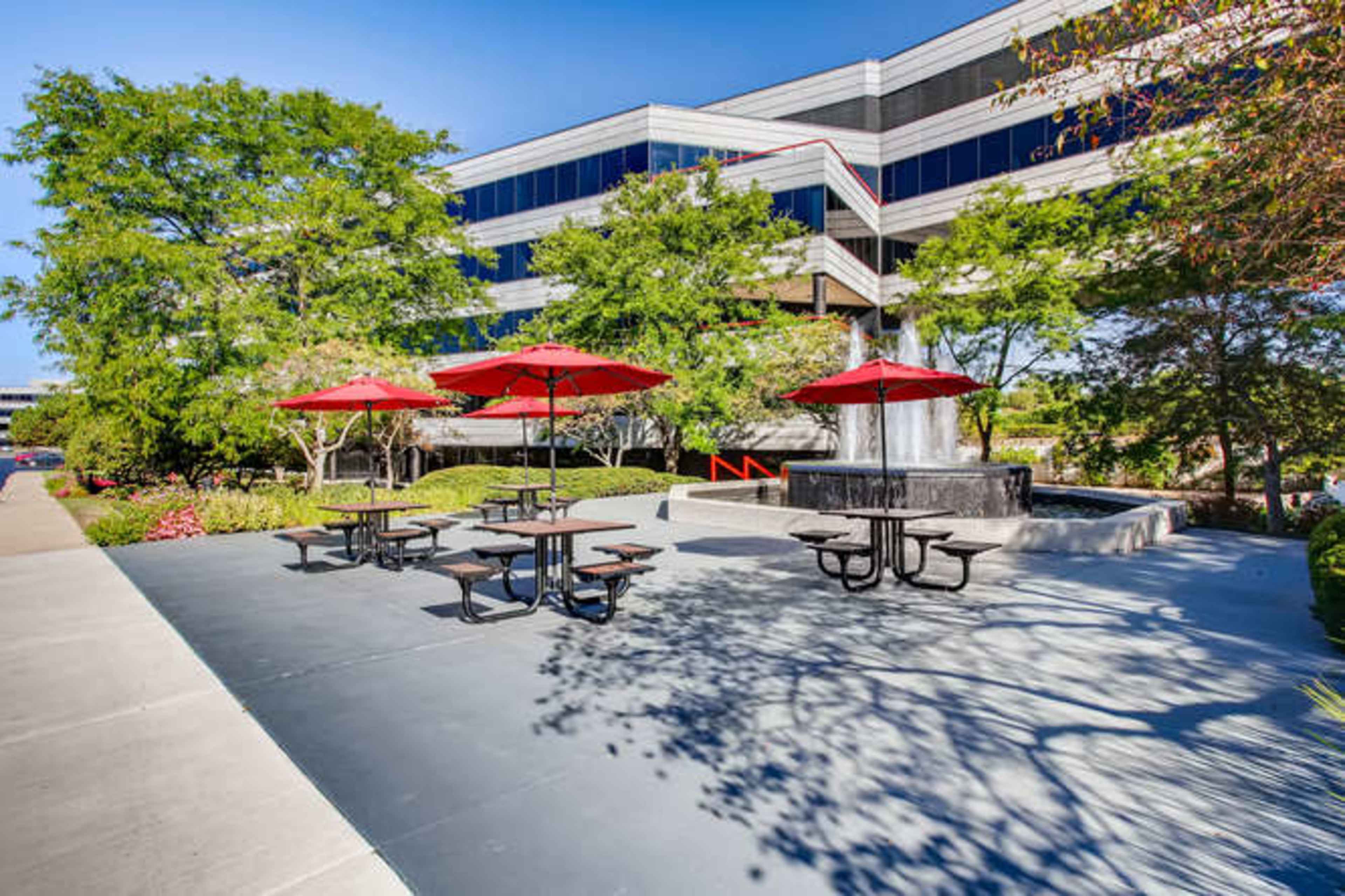 The image shows an outdoor seating area with picnic tables and red umbrellas near a multi-story office building, surrounded by greenery.