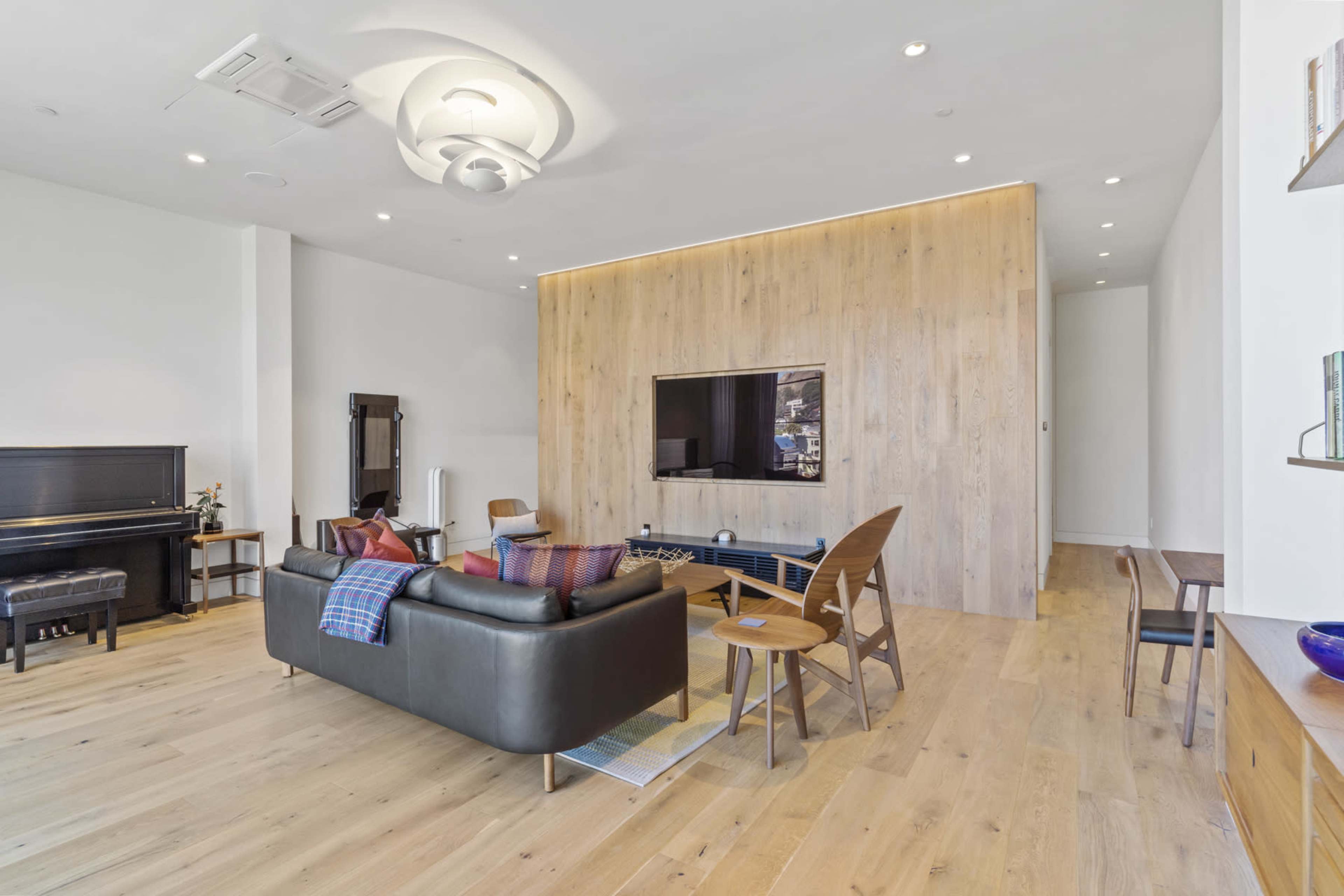 The image shows a spacious living room featuring a wooden accent wall, a large TV, a piano, and various seating arrangements with simple decor.