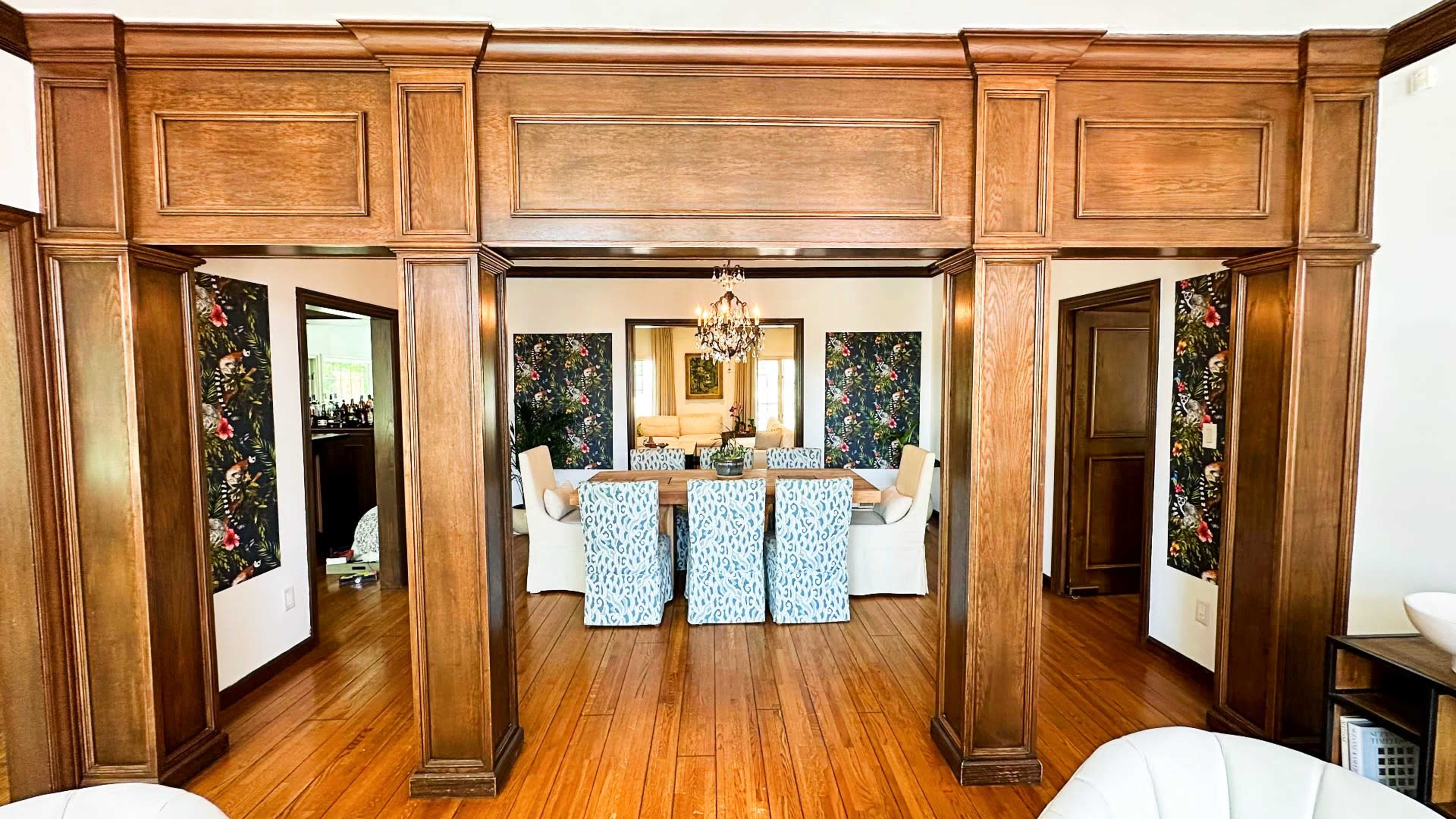 The image shows a spacious interior with wooden columns framing a view into a dining and living area, decorated with floral artwork.