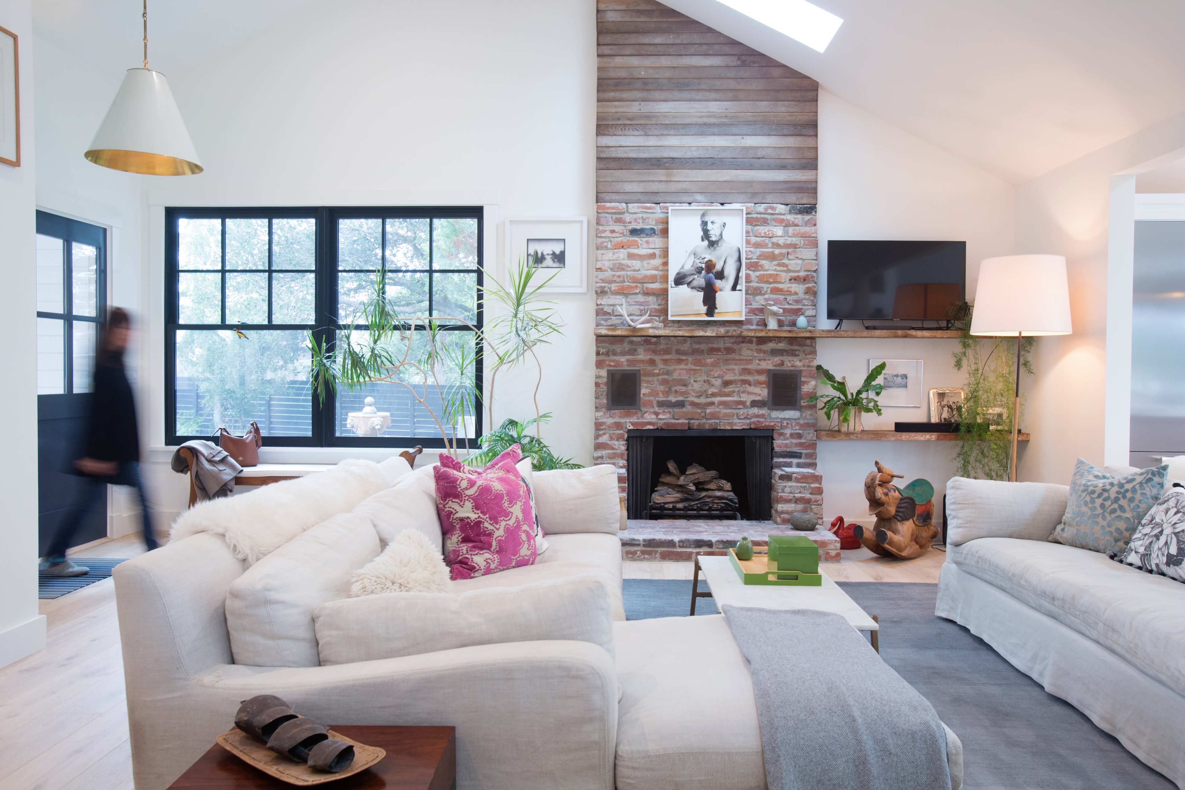 A modern living room with two large sofas, a brick fireplace, plants, and large windows allowing natural light.