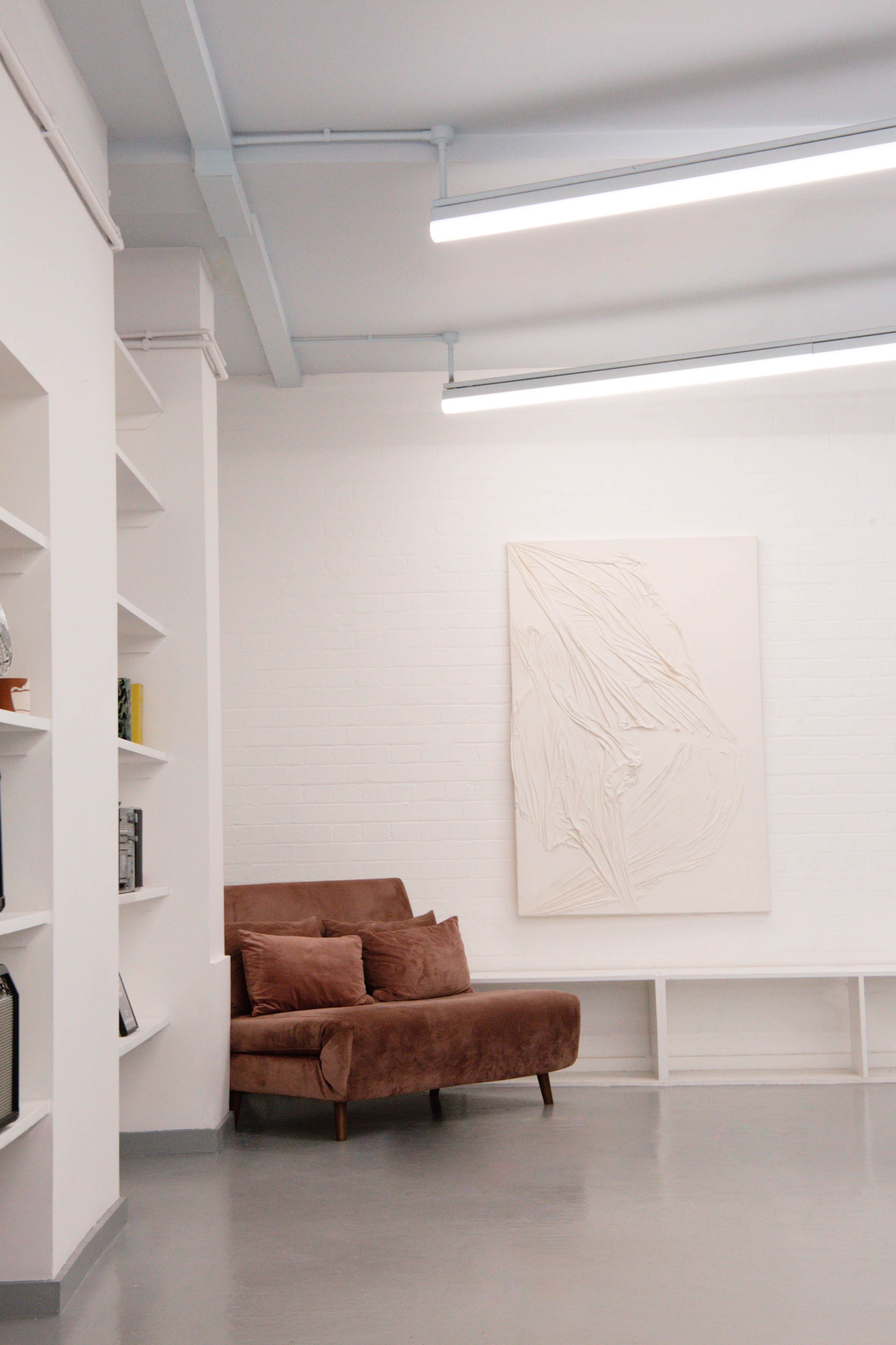The image shows a modern interior with a brown velvet sofa, a white textured wall art piece, and shelves on the left side displaying various items.