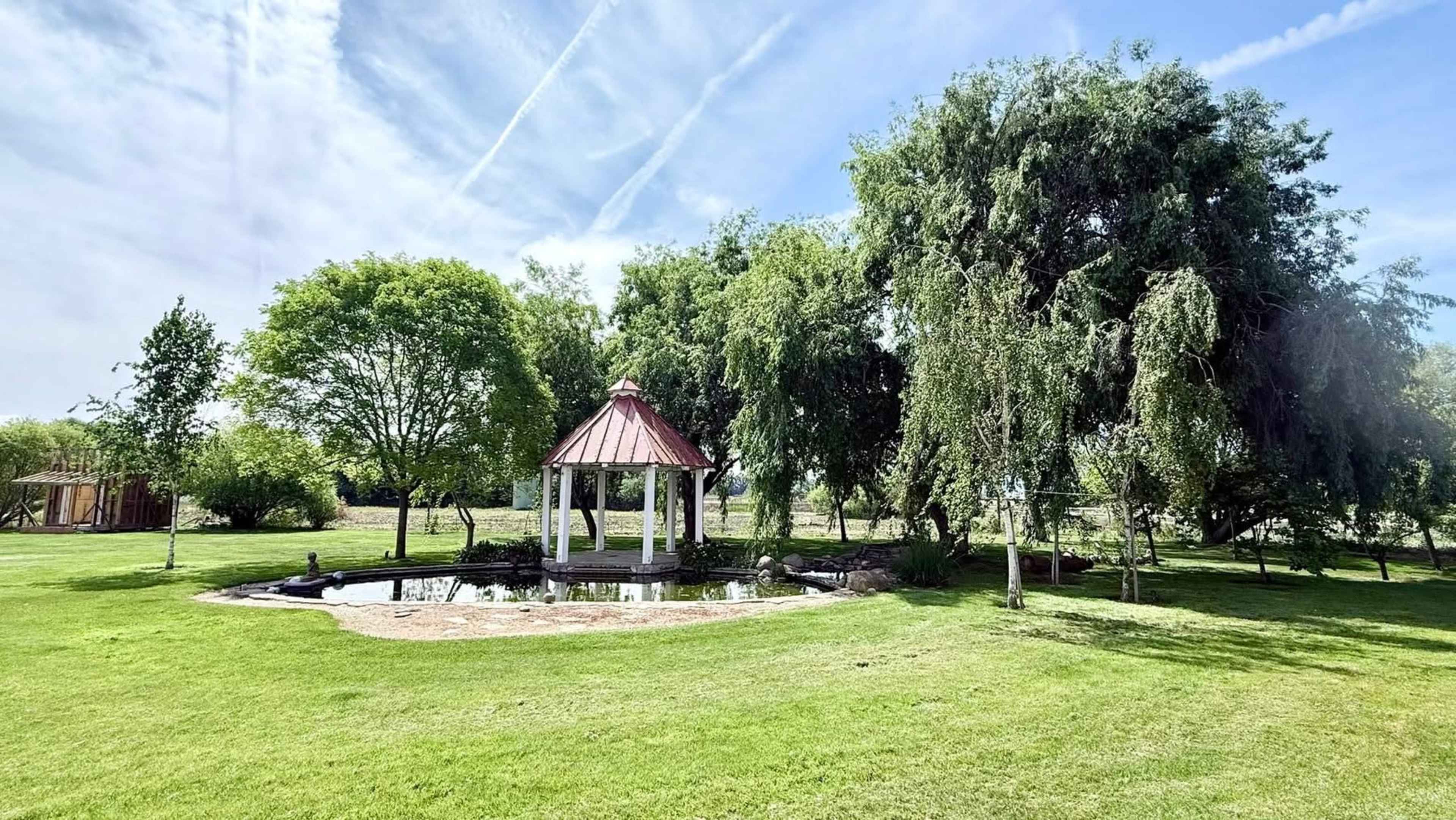 A gazebo with a red roof stands near a pond, surrounded by greenery and trees in a park.