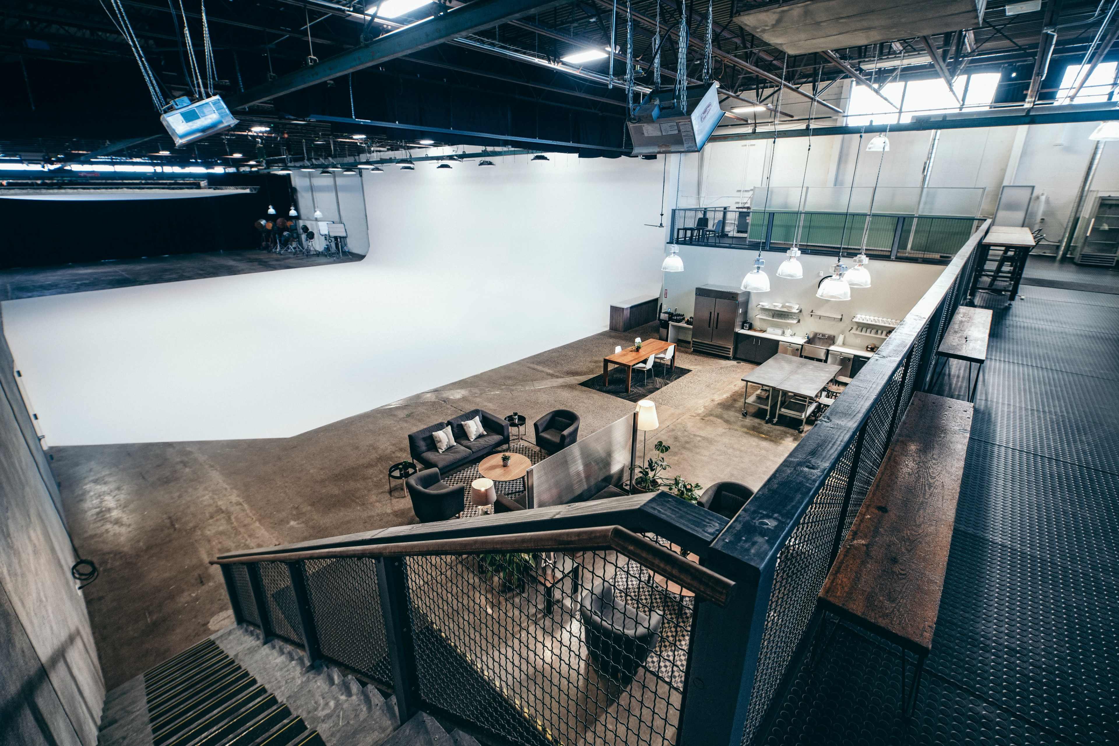 The image shows a spacious studio with a large, empty white backdrop, various seating arrangements, and tables in an industrial-style setting.