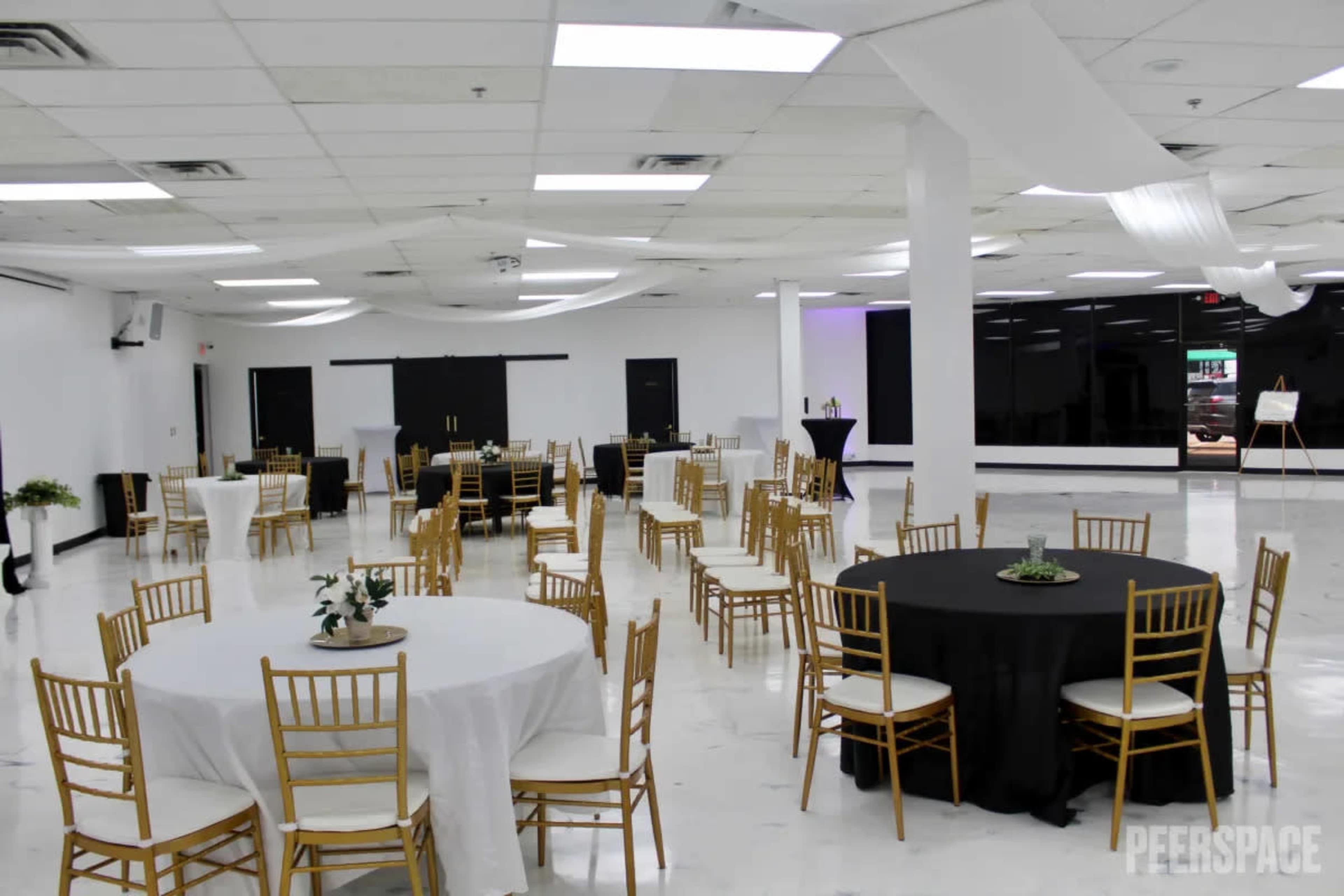 A spacious event hall features several round tables with gold chairs, evenly distributed across a glossy white floor under soft lighting.