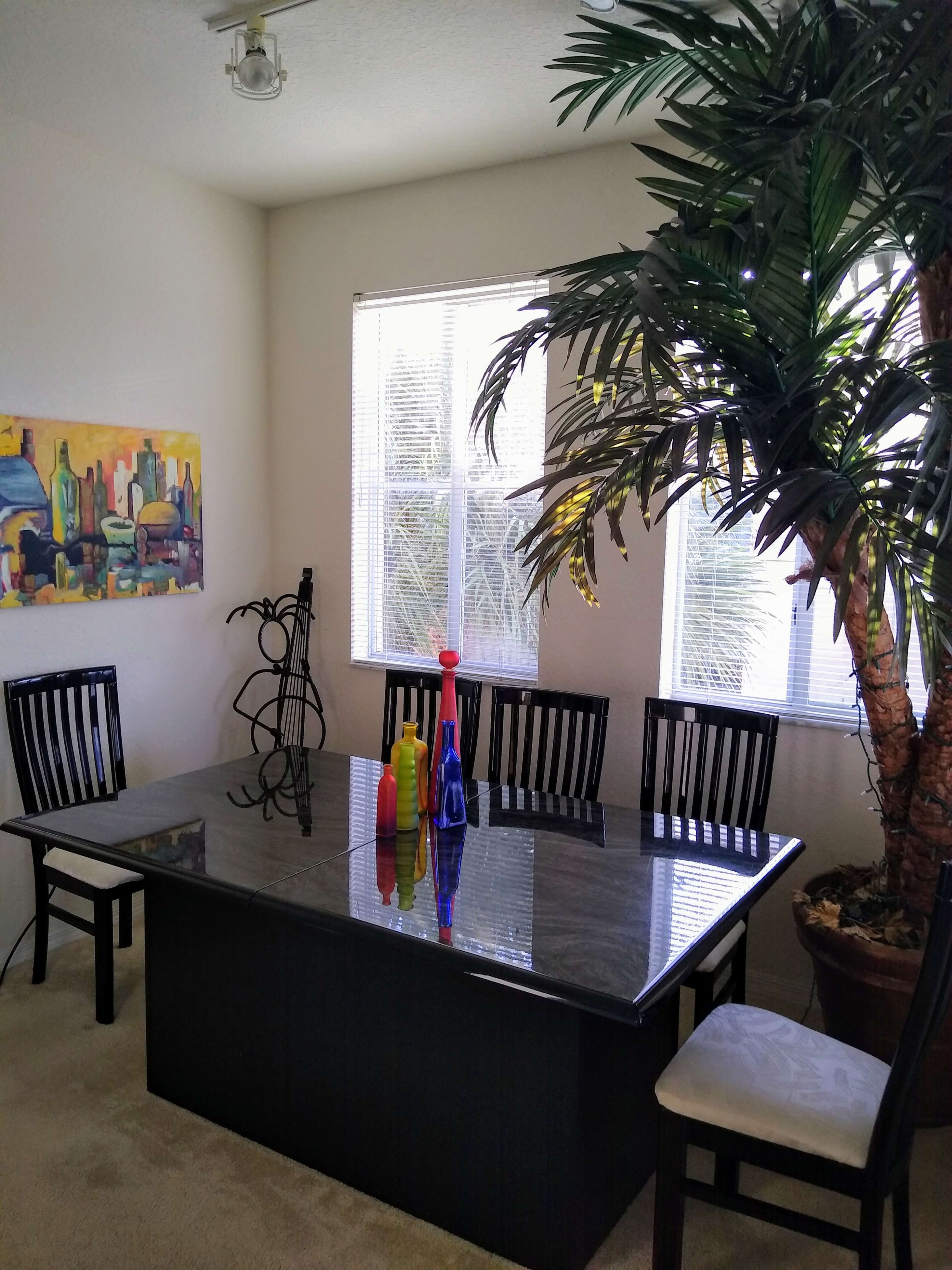 A dining room features a large black table surrounded by six wooden chairs, with colorful bottles in the center and a potted palm tree in the corner.