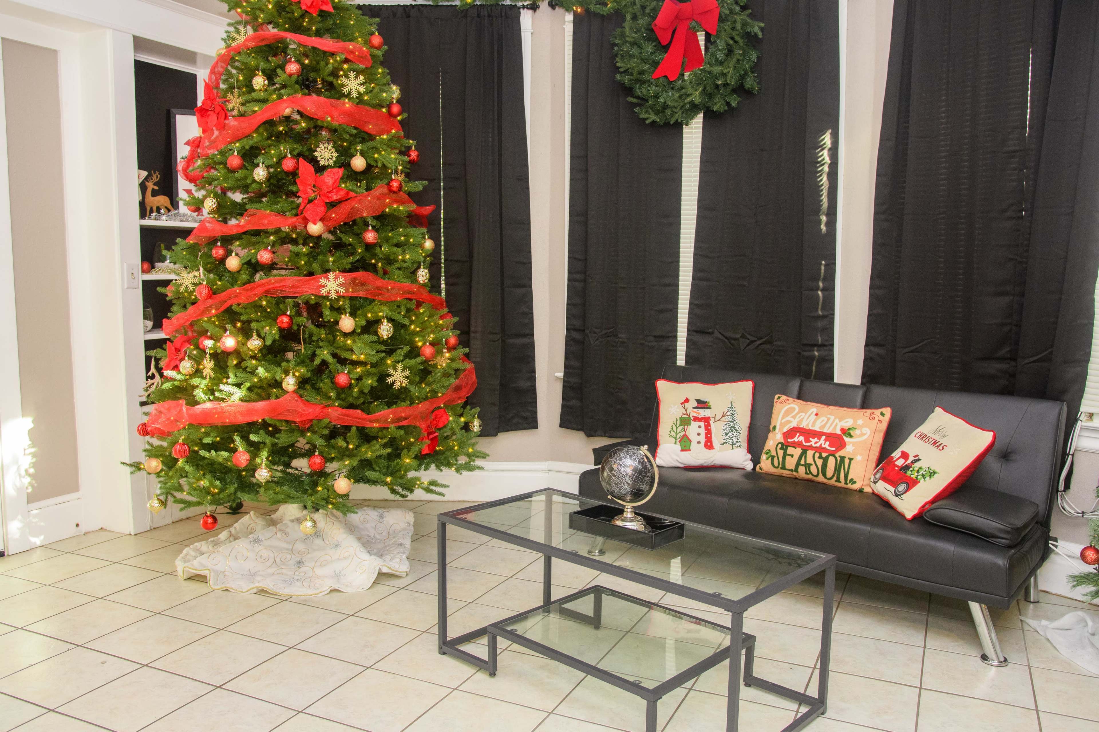 The scene features a decorated Christmas tree with red ribbons and ornaments, positioned next to a black sofa adorned with festive throw pillows, all set in a room with black curtains and a glass coffee table.