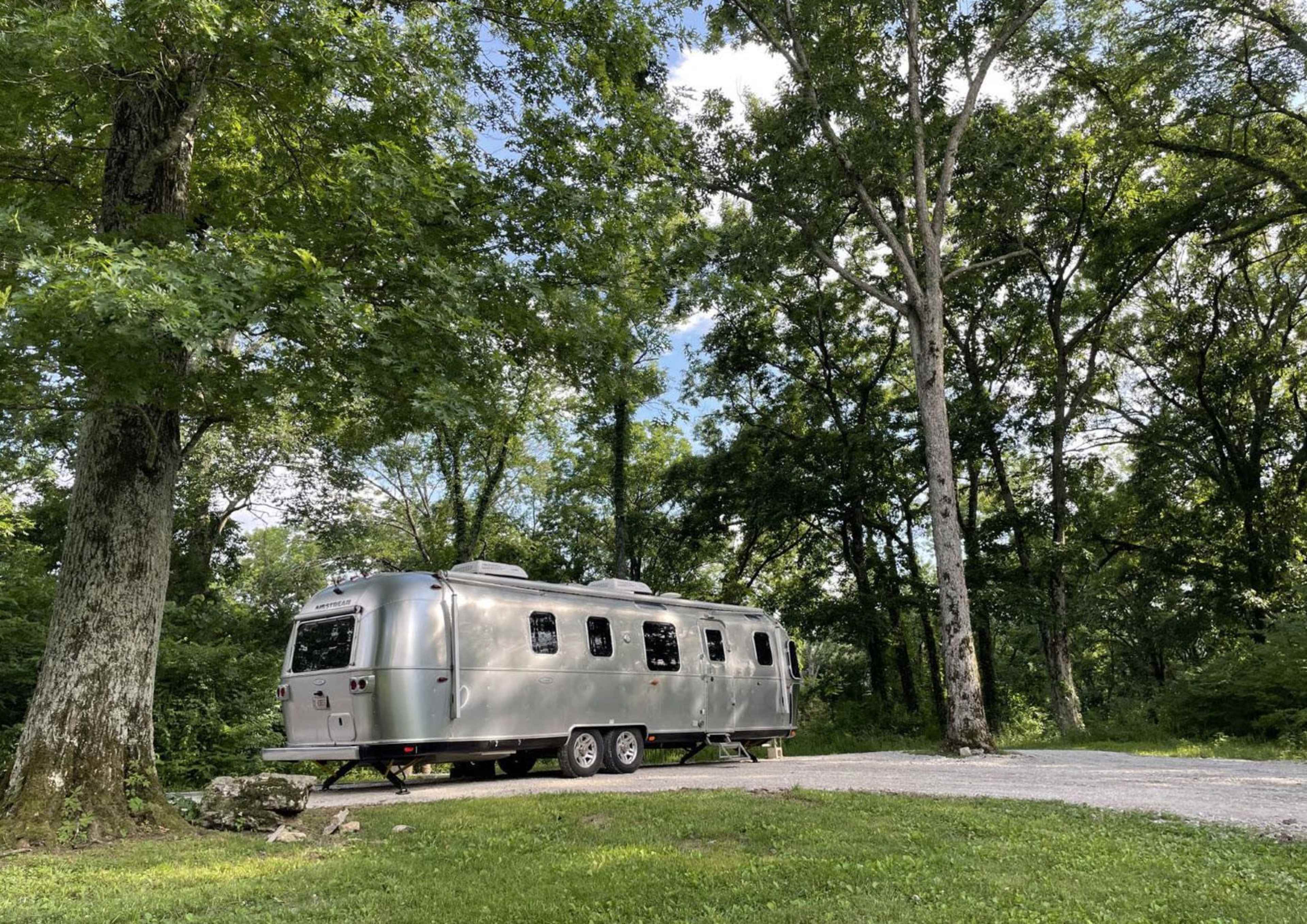 A silver Airstream trailer is parked on a gravel site surrounded by tall green trees.