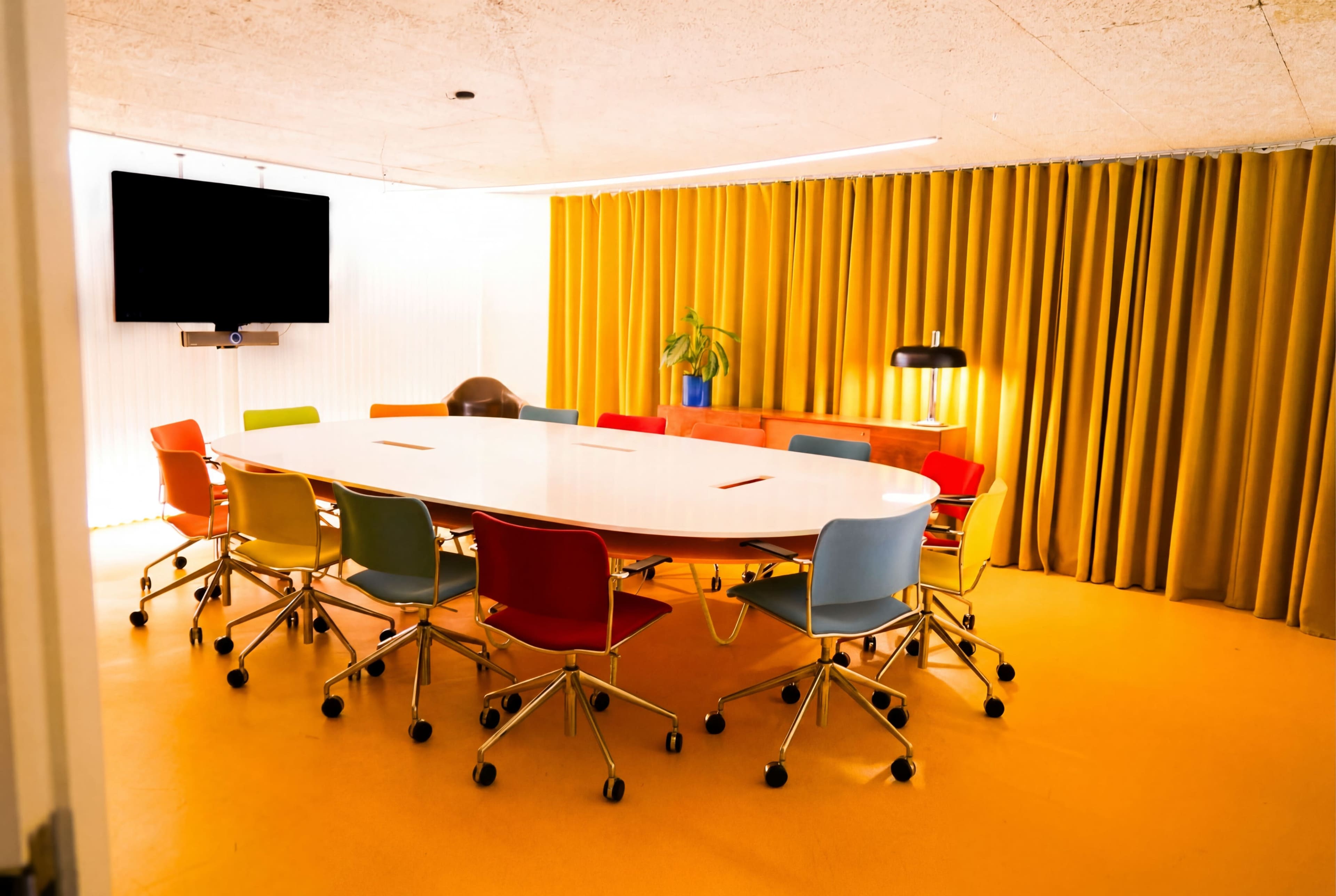 A bright and modern conference room features a large oval table surrounded by colorful chairs, with a television and large yellow curtains in the background.