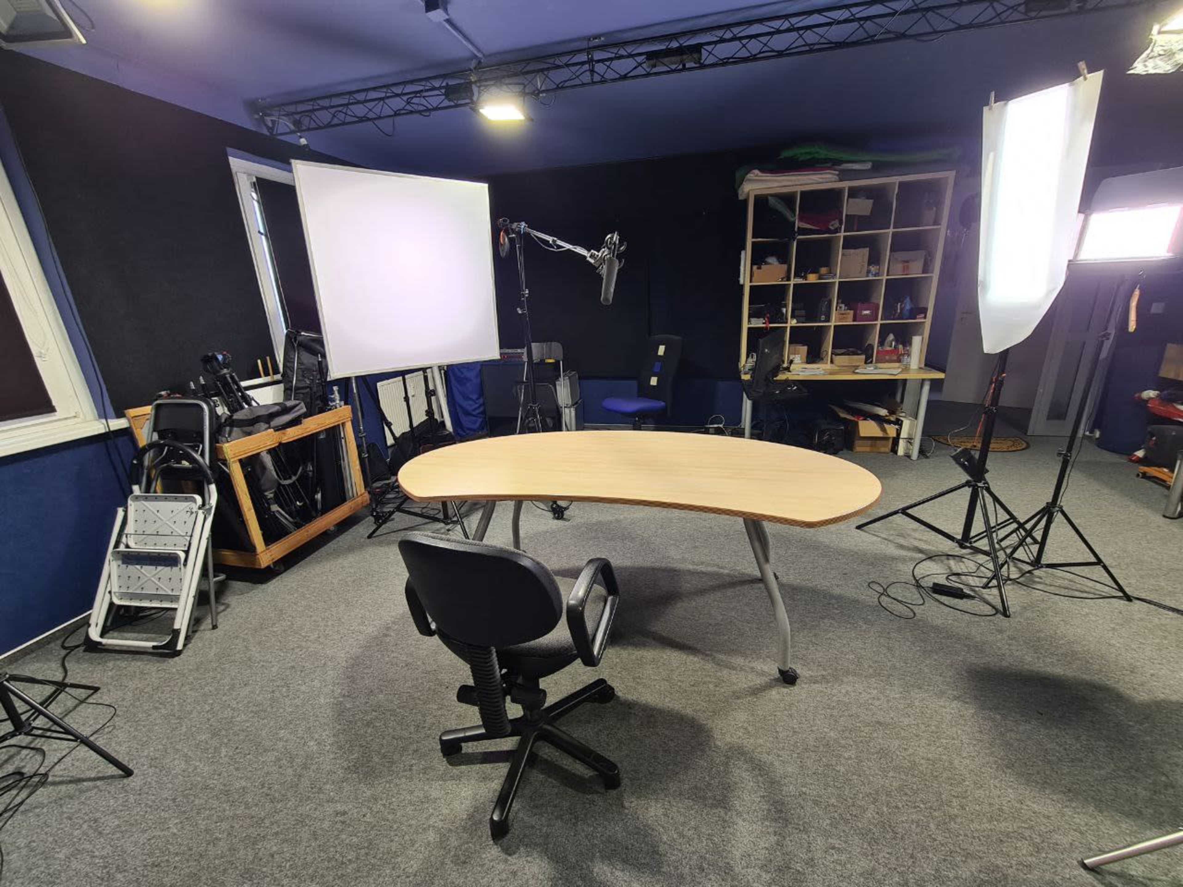 A studio setup features a wooden oval table and an office chair surrounded by various lighting equipment and a microphone.