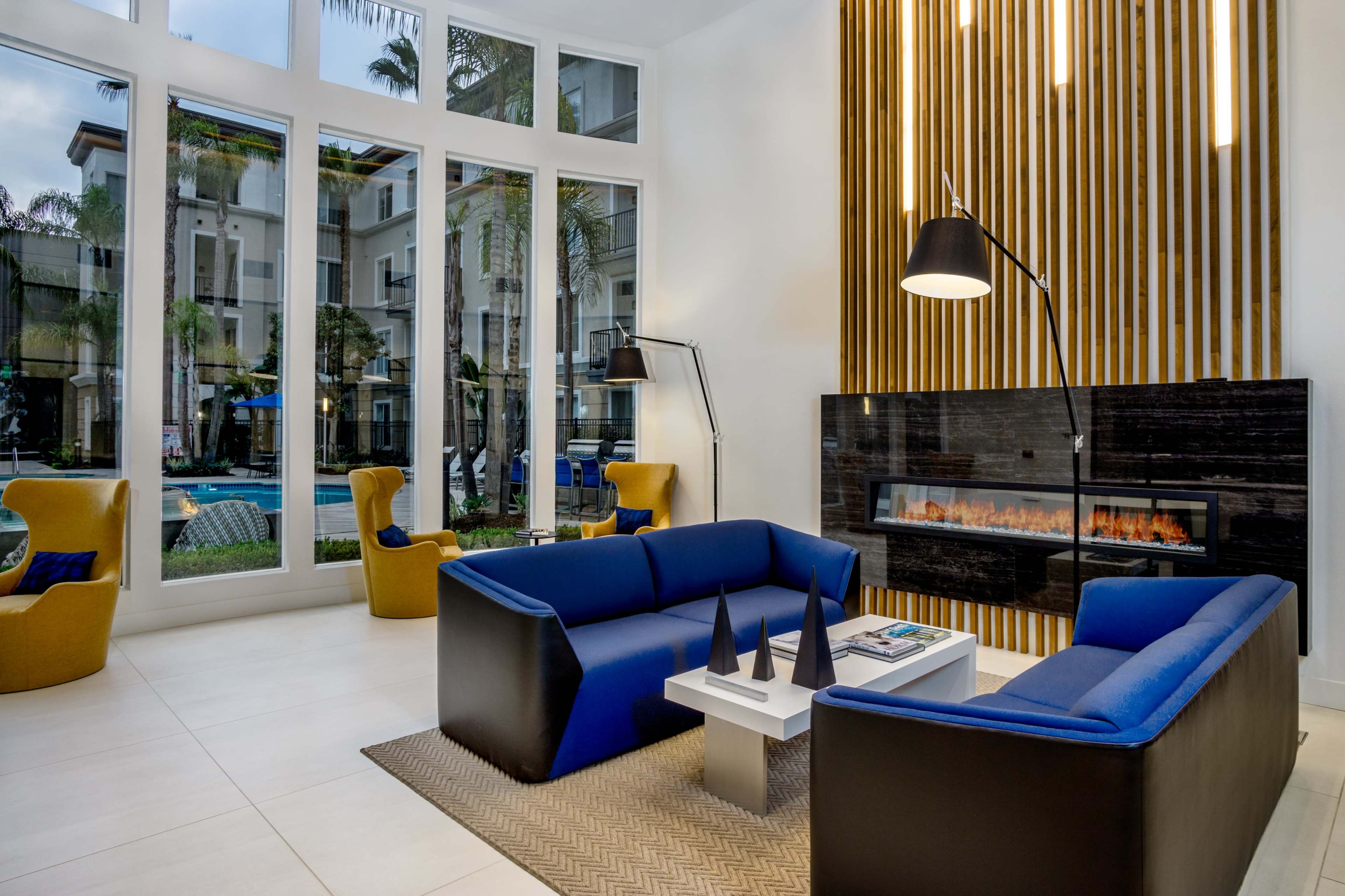 A modern lounge area with large windows, featuring two blue sofas, a coffee table, and a black fireplace against a wood-paneled wall.