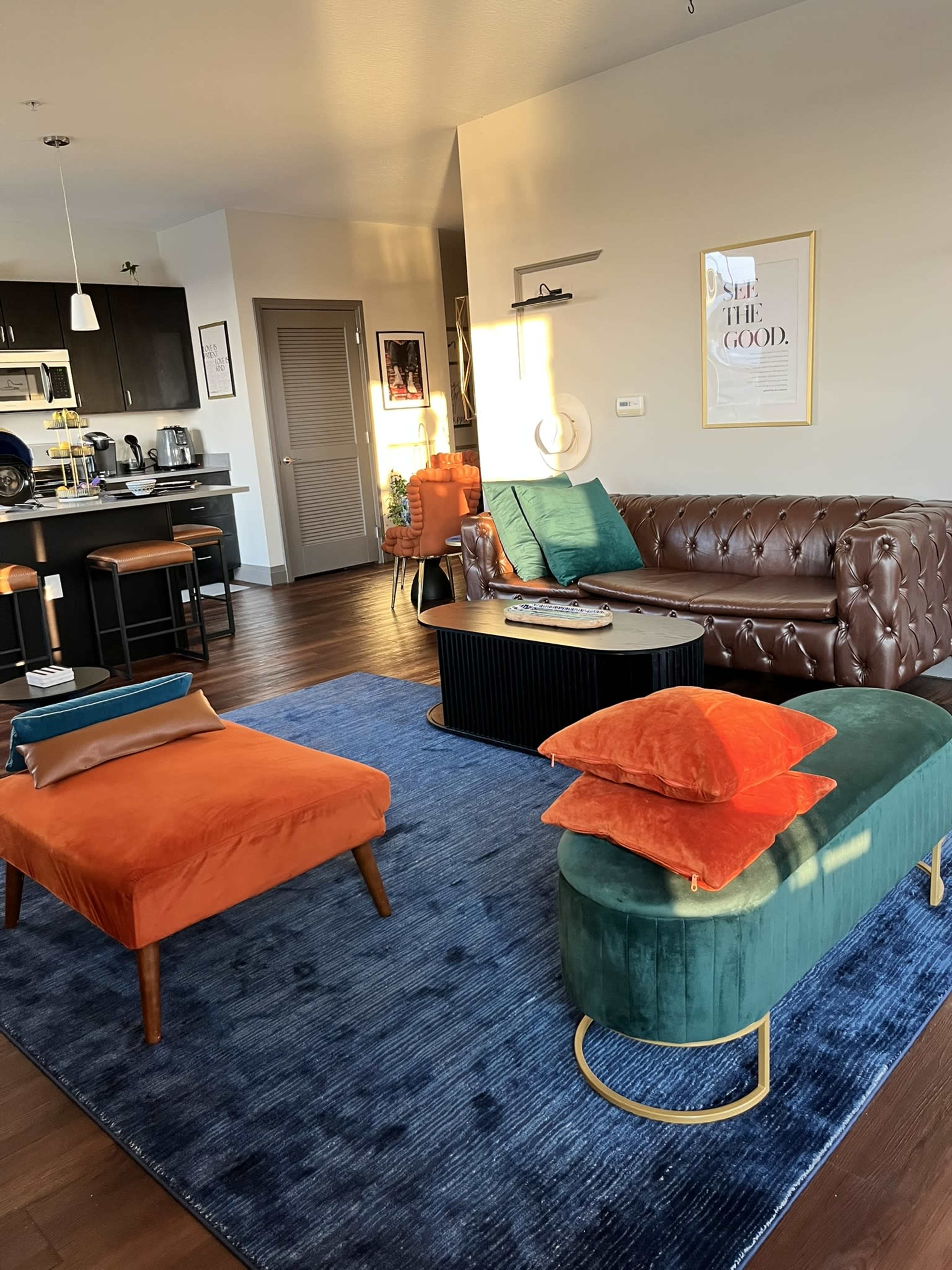 The image shows a modern living room with a brown leather sofa, colorful ottomans, a black coffee table, and a kitchen area in the background.