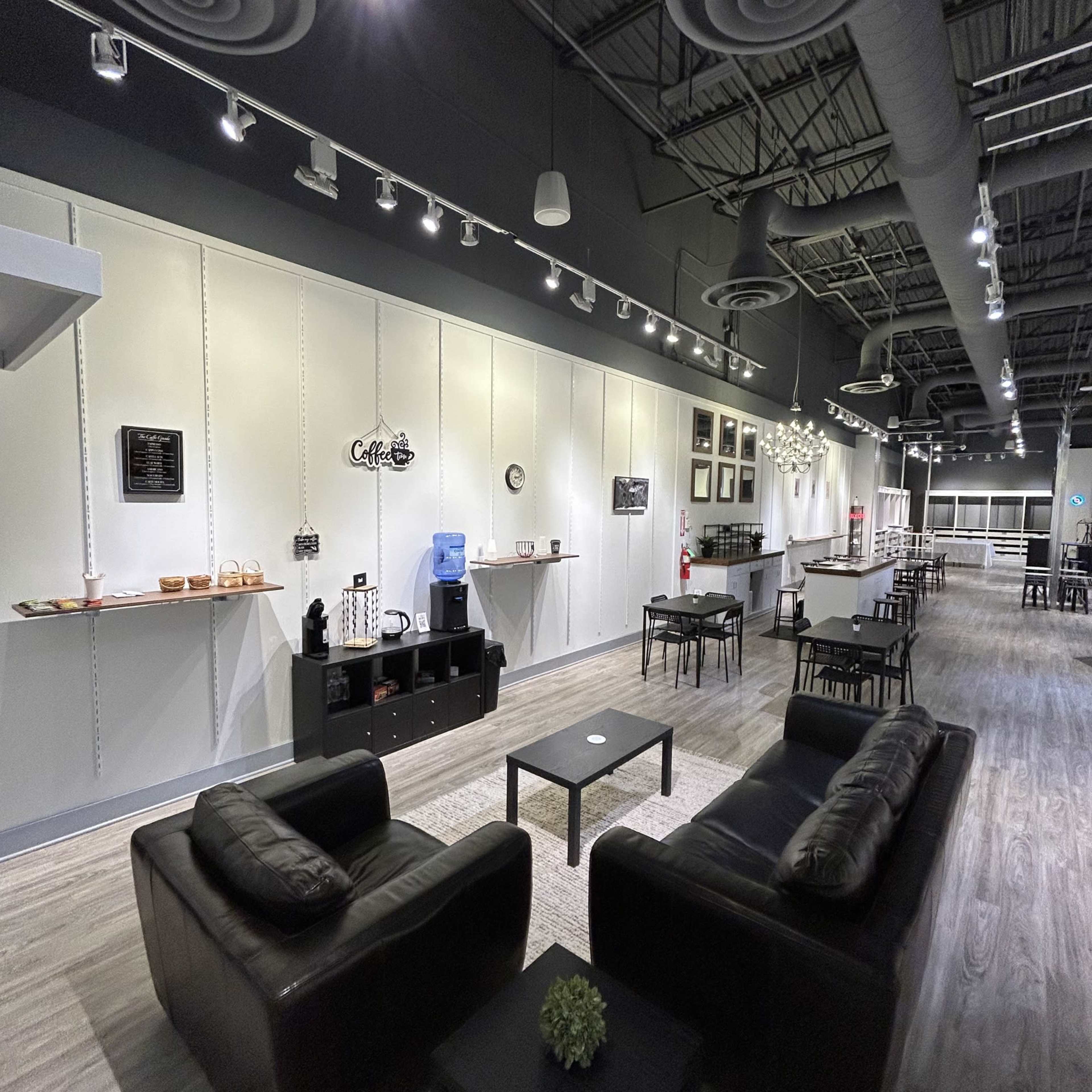 The image shows a modern lounge area with black leather couches, a coffee station, and various decorative wall shelves in a spacious room.