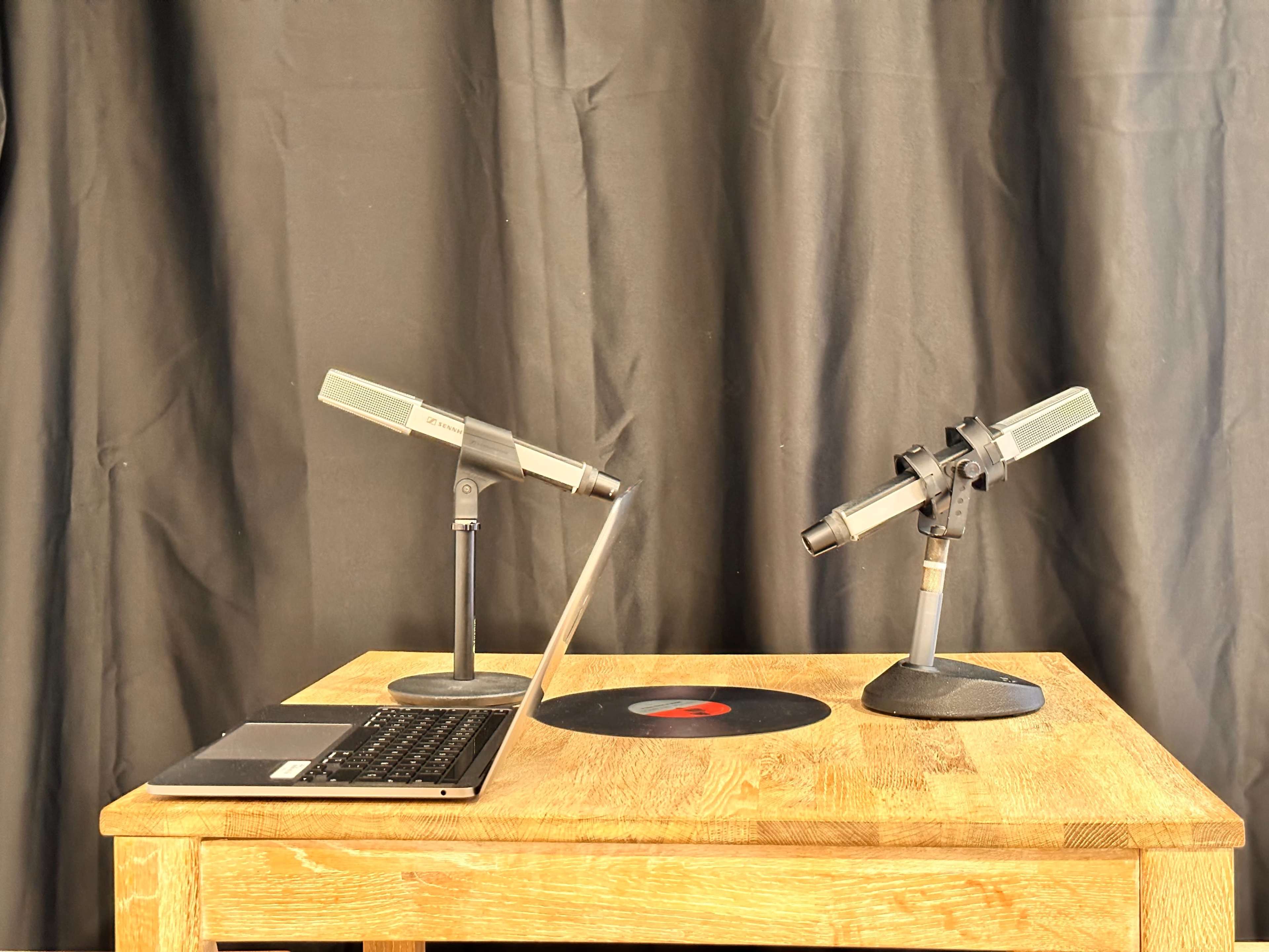 A wooden table displays two microphones on stands and a laptop next to a vinyl record against a black backdrop.