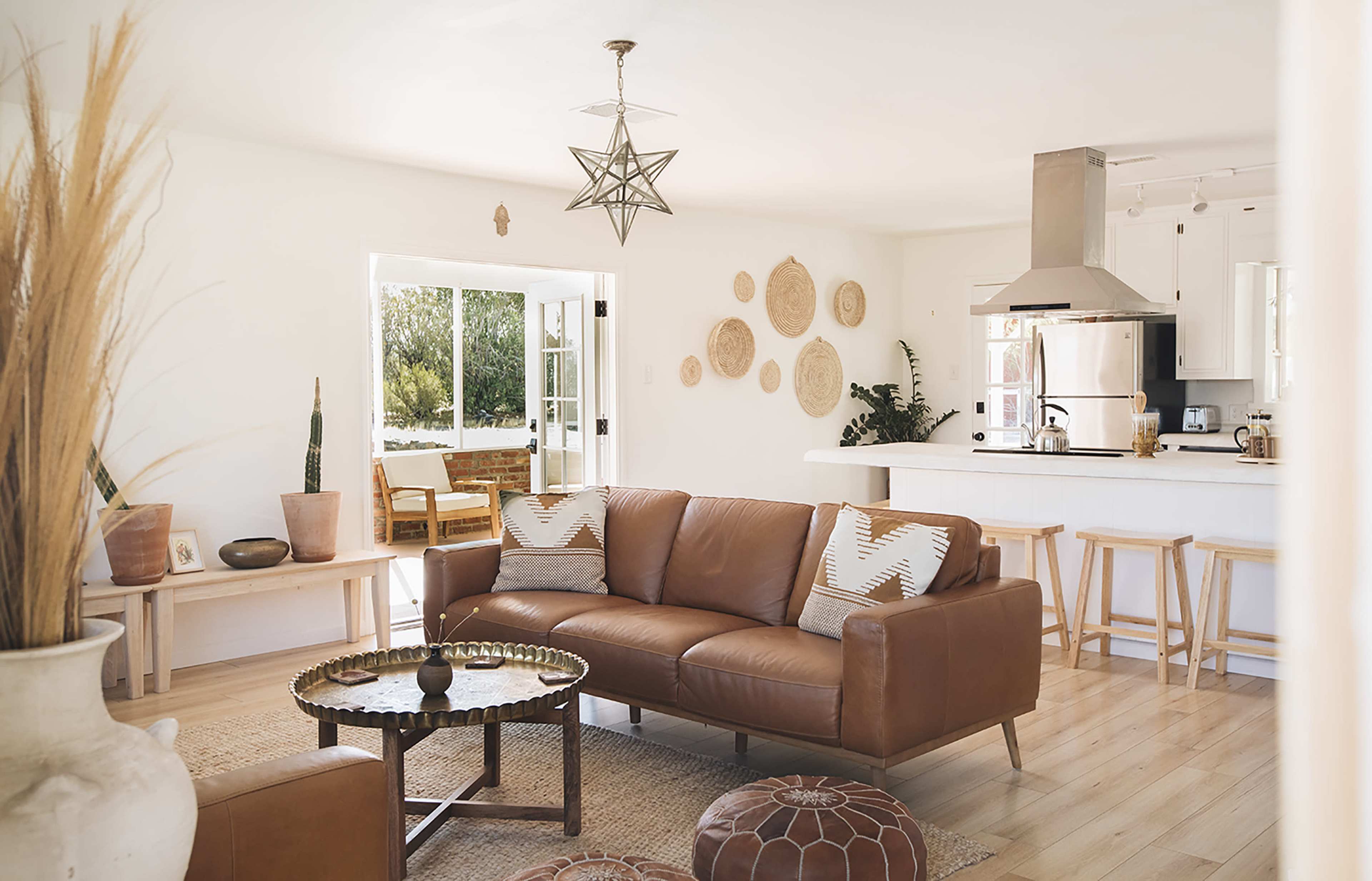 The image shows a modern living room with a brown leather sofa, a round coffee table, and a dining area visible through an open door, accented by plants and decorative wall hangings.