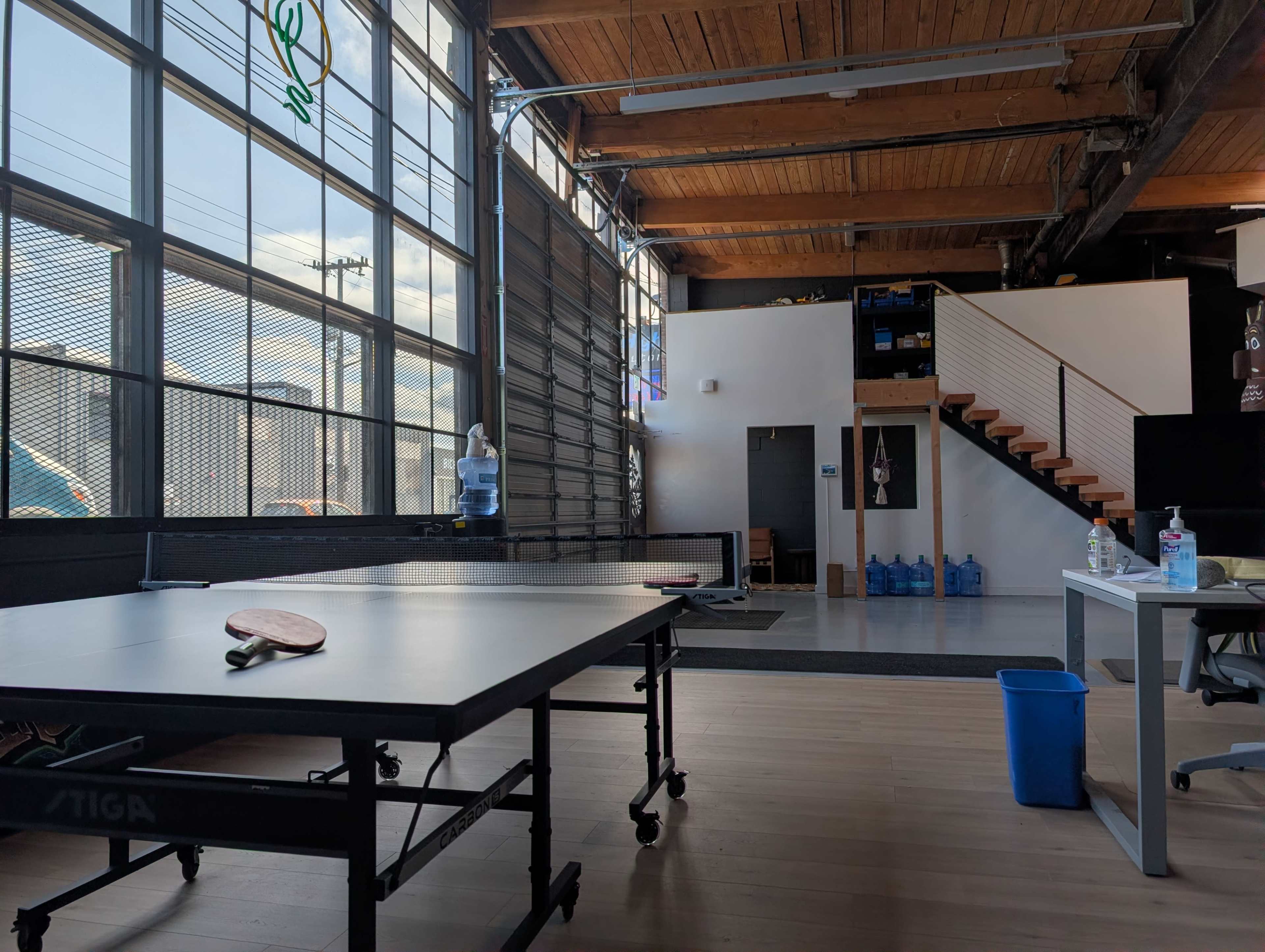 The image shows a modern, spacious room with large windows, featuring a table tennis table, a water cooler, and stairs leading to an upper level.