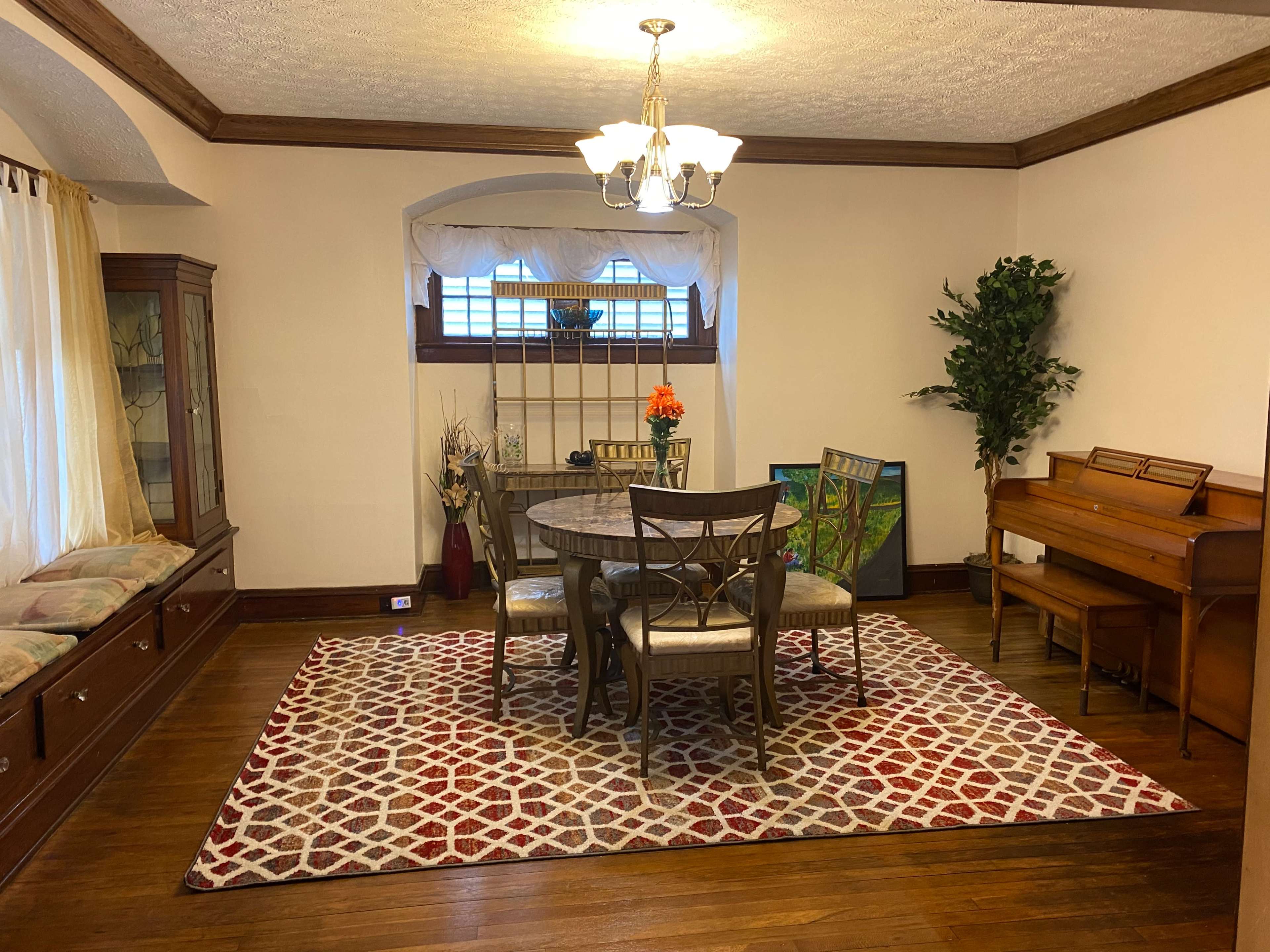 A dining area with a round table and four chairs, surrounded by a patterned rug, a wooden piano, and a display cabinet.