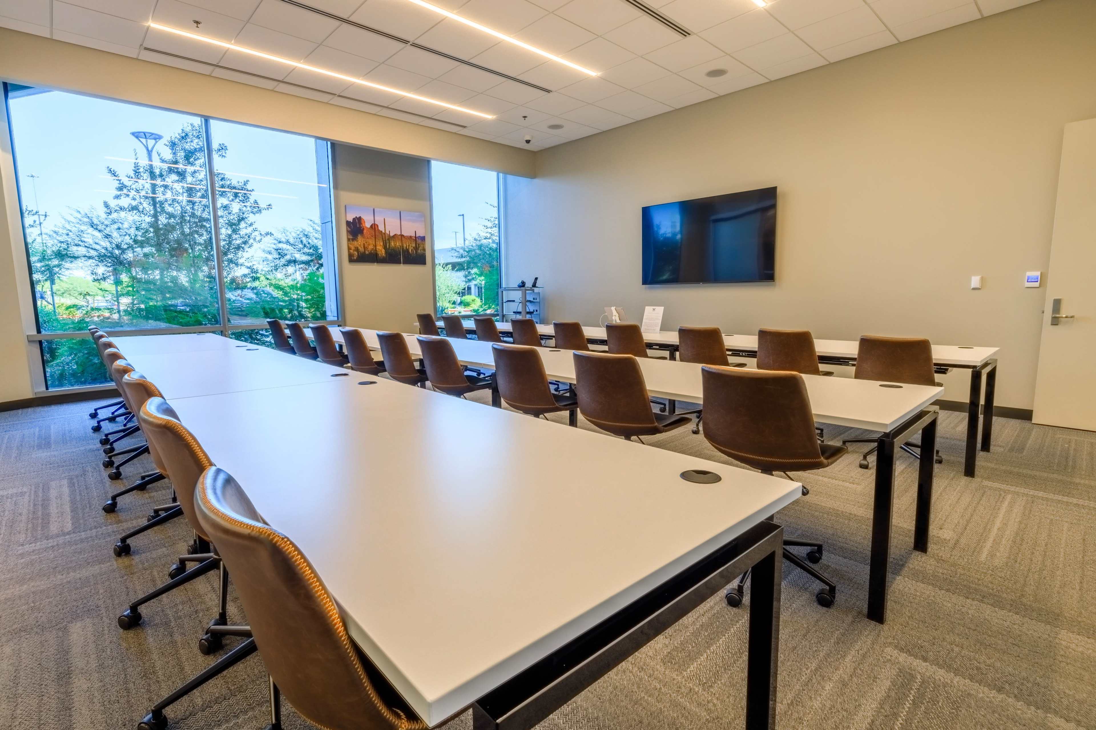 A modern conference room features rows of tables and chairs facing a large screen, with natural light streaming in through a window.