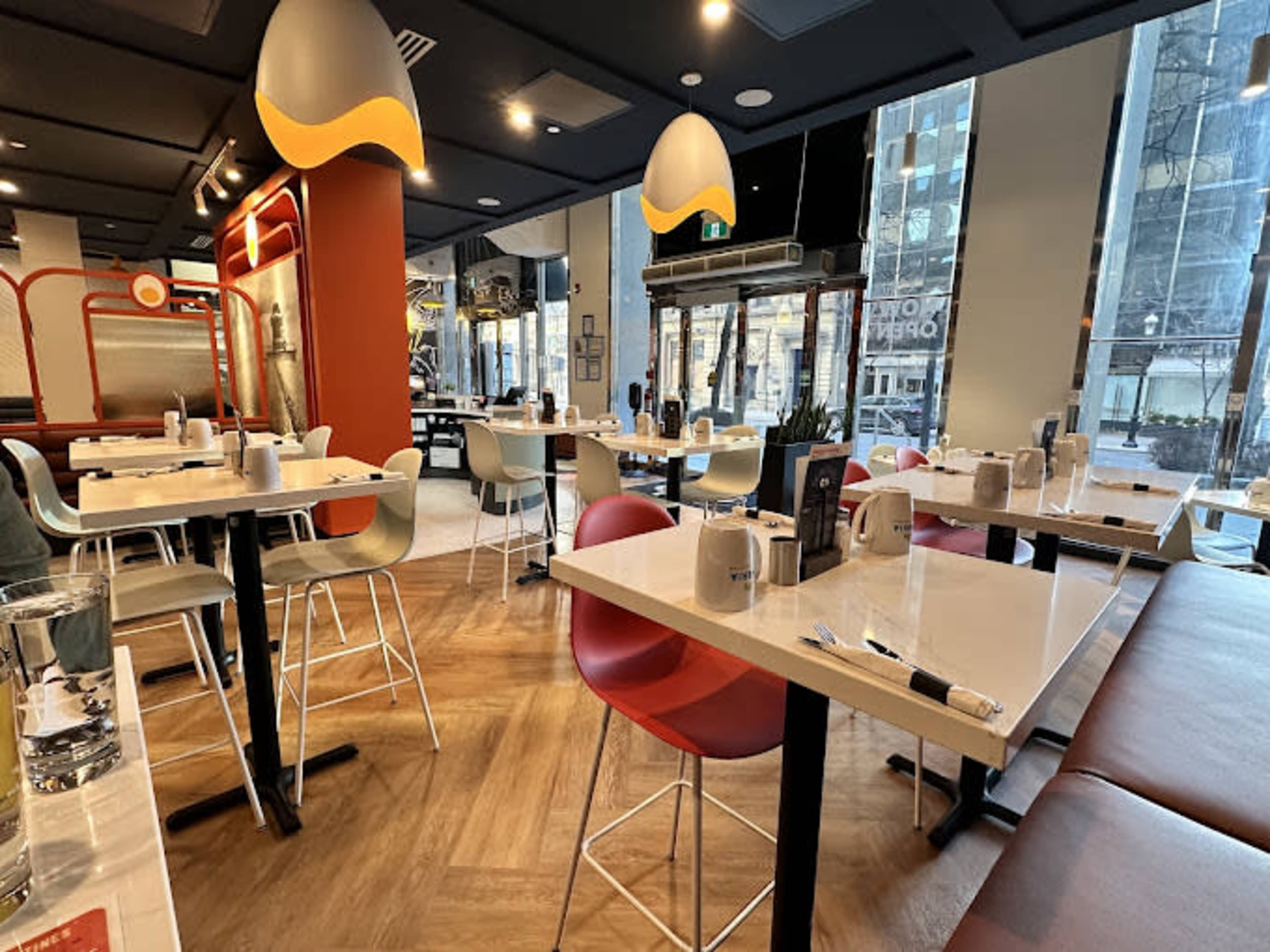 The image shows a modern restaurant interior with several tables set for dining, featuring colorful chairs and large windows allowing natural light to enter.