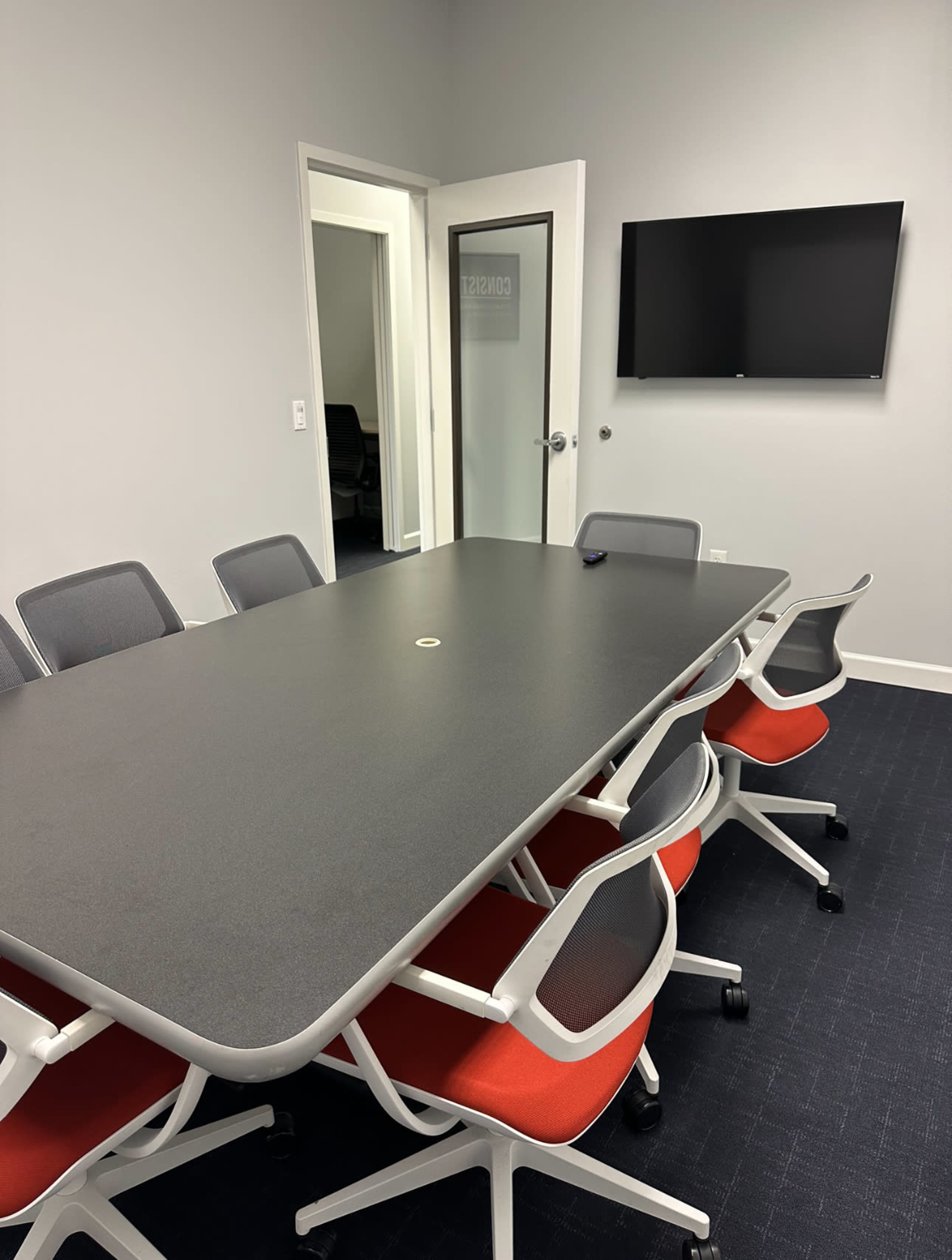 The image shows a modern conference room with a large table surrounded by eight chairs and a wall-mounted TV.
